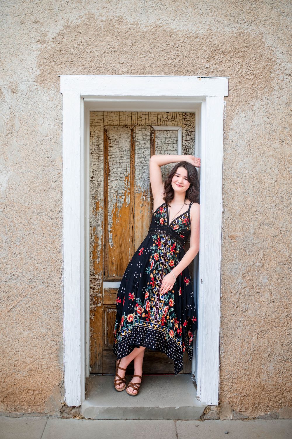 A young woman in a floral maxi dress poses gracefully in a vintage white doorframe against a stucco wall.