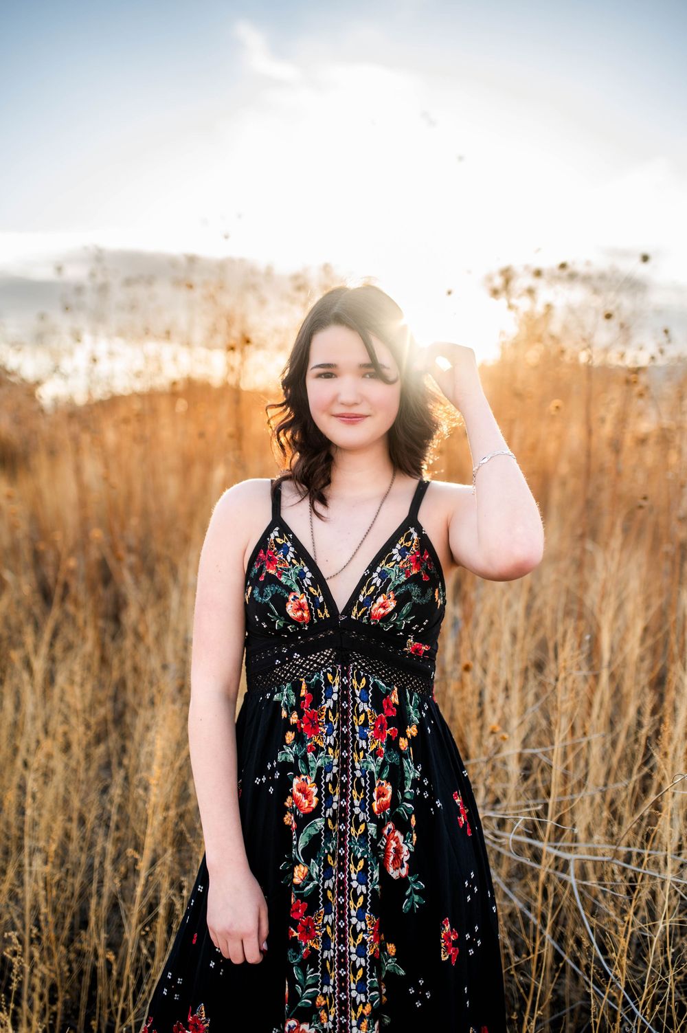 A young woman in a black floral dress stands in a golden field at sunset with mountains behind her.