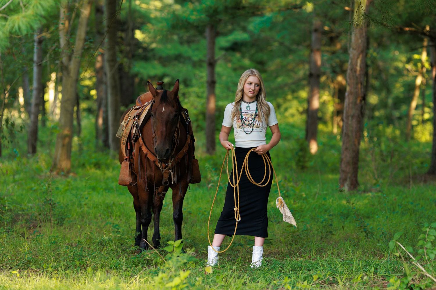 High school senior girl posing fiercely with her horse while holding loops of her rope, wearing a black skirt, graphic T and turquoise.