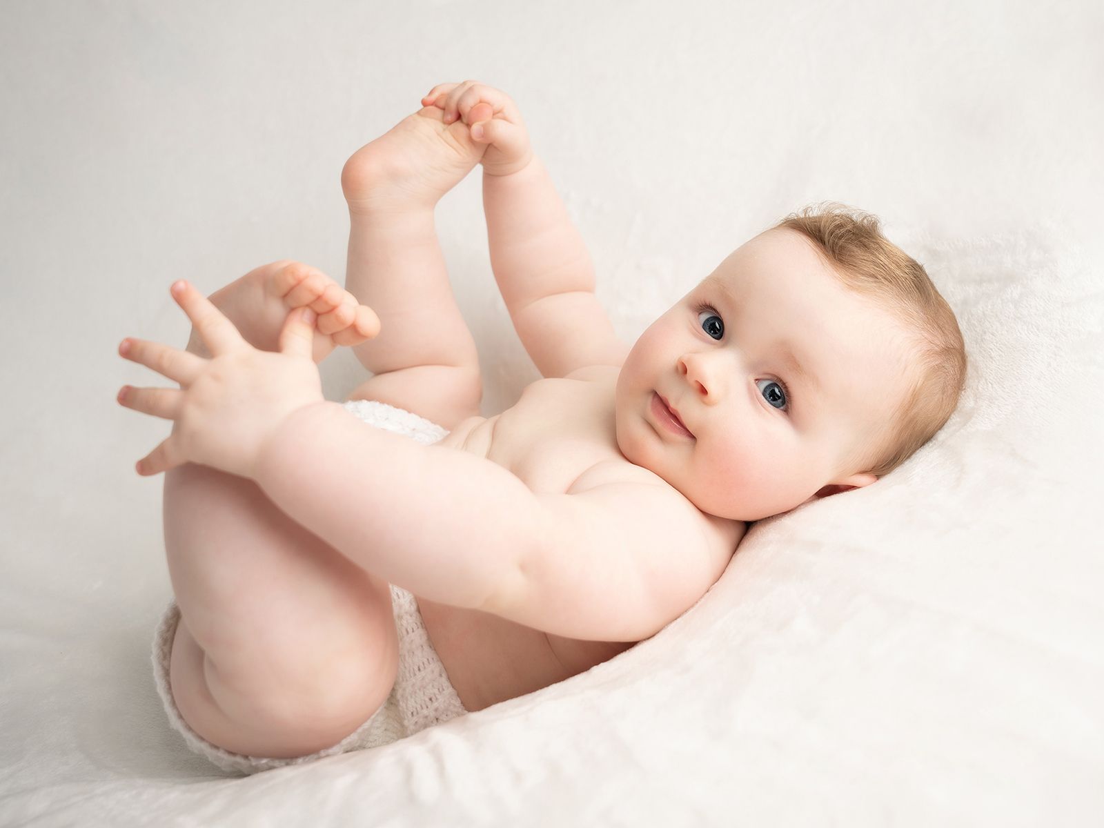 Playful baby in white outfit holding feet while lying on blanket.