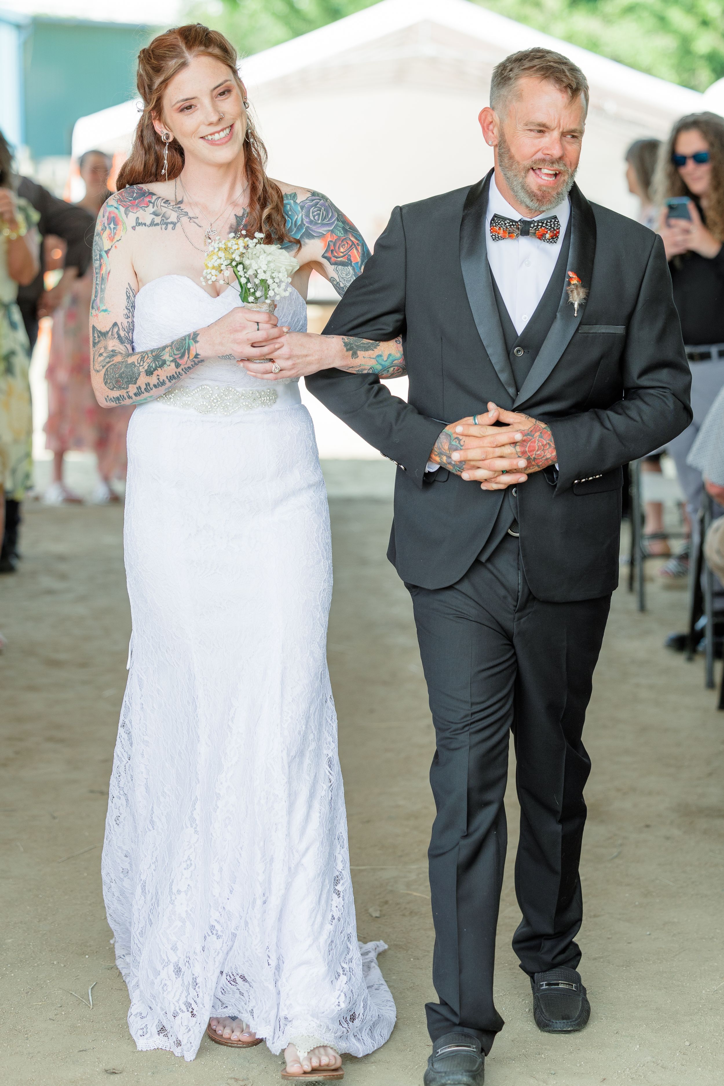 Smiling bride with sleeve tattoos in white lace gown walking down aisle with tattooed companion.