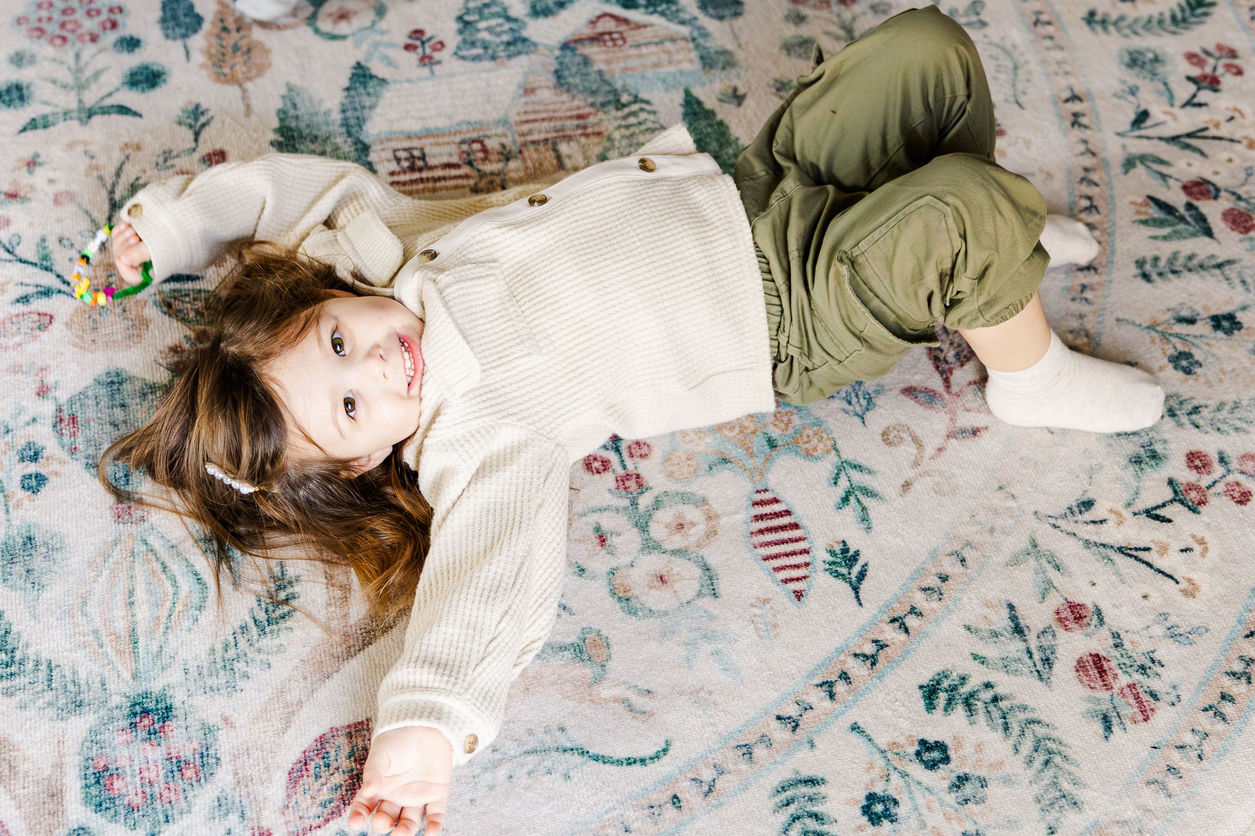 Adorable young girl lying on floral patterned rug looking up at camera with a smile.