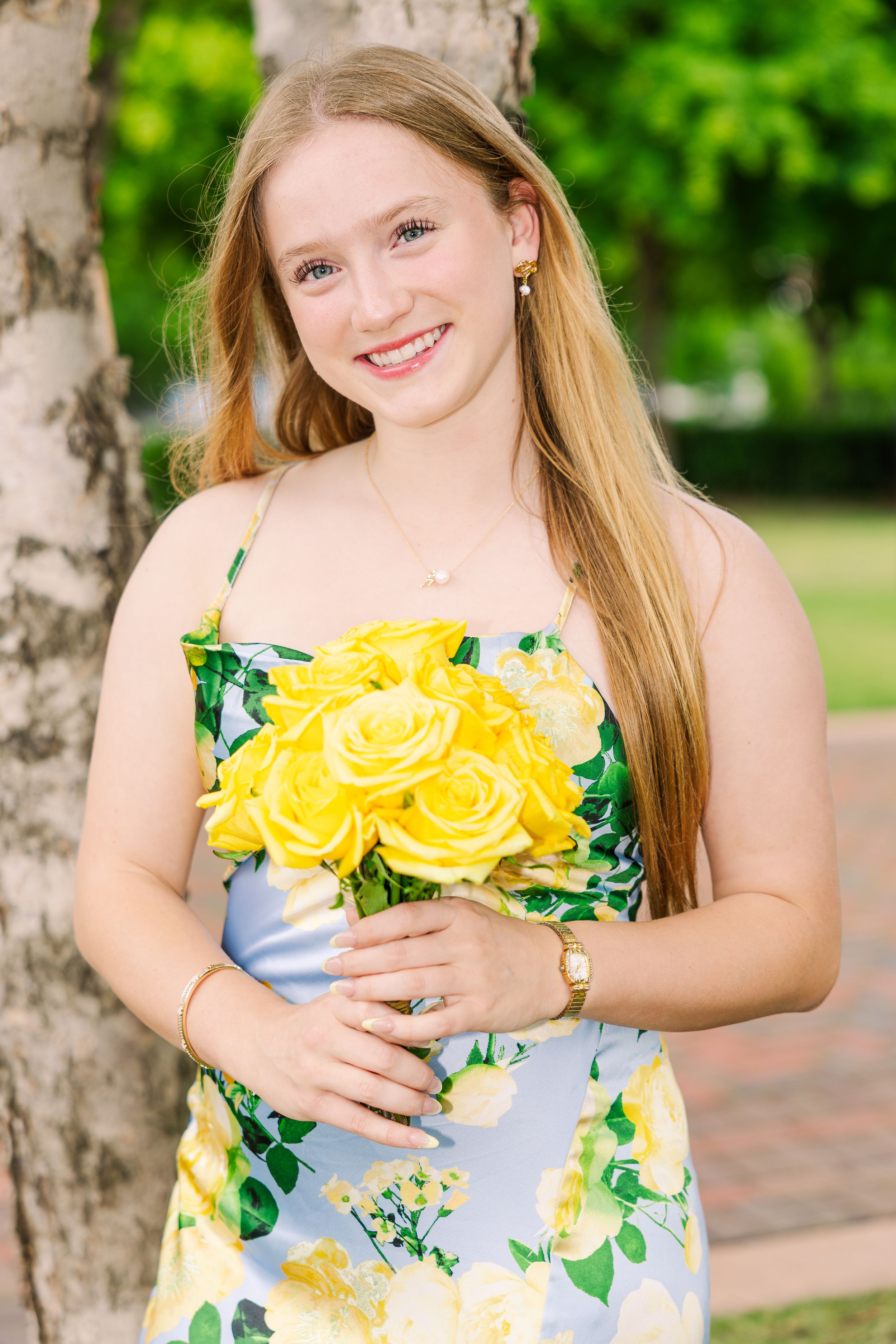 Smiling young woman in floral yellow dress holding bouquet of yellow roses outdoors.