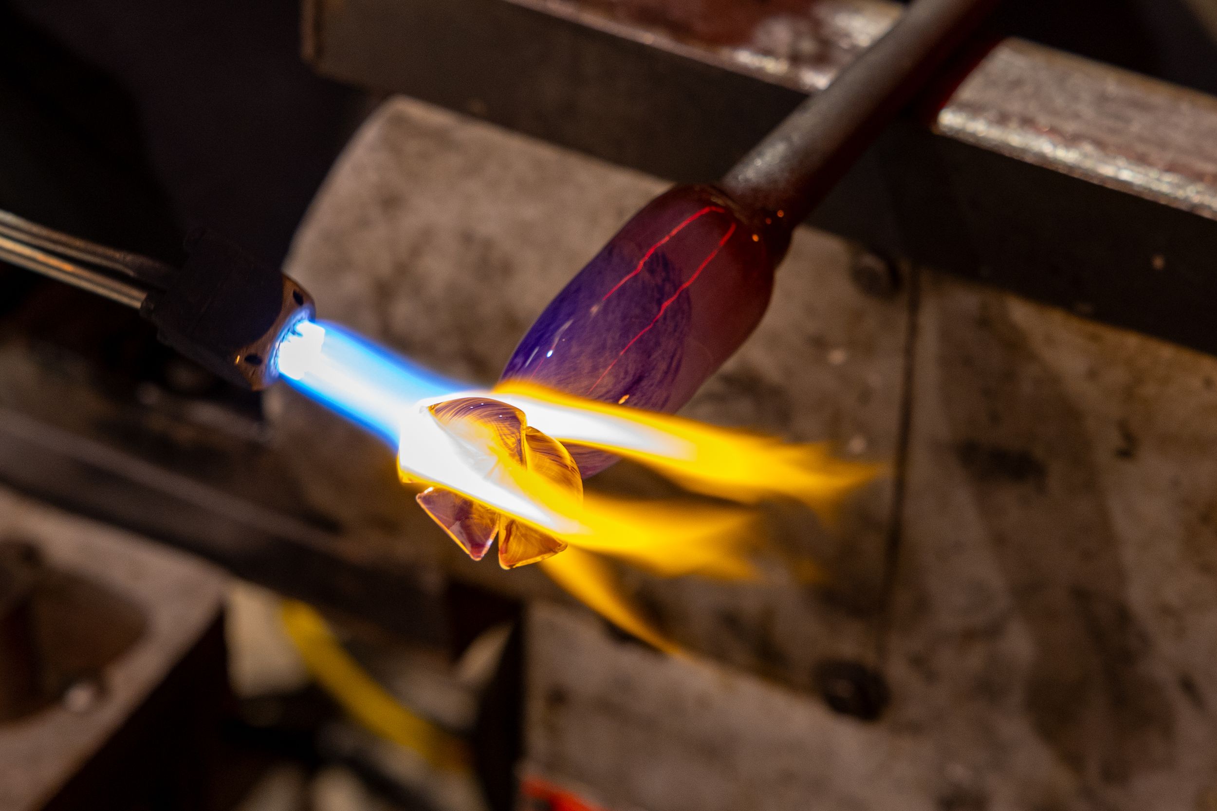 A glassblower uses a torch flame to heat colorful molten glass in a workshop.