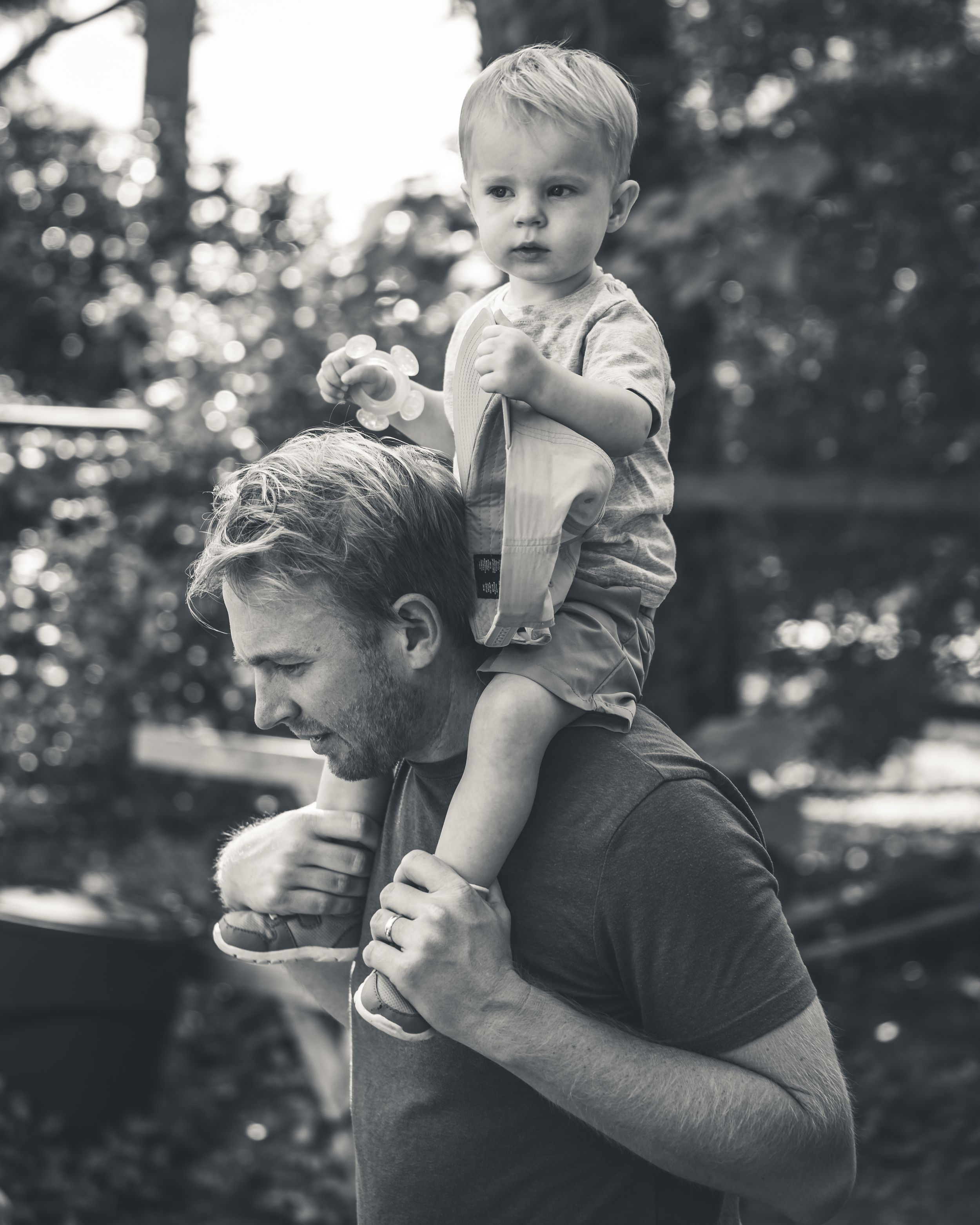 Black and white photo of toddler sitting on father's shoulders outdoors.