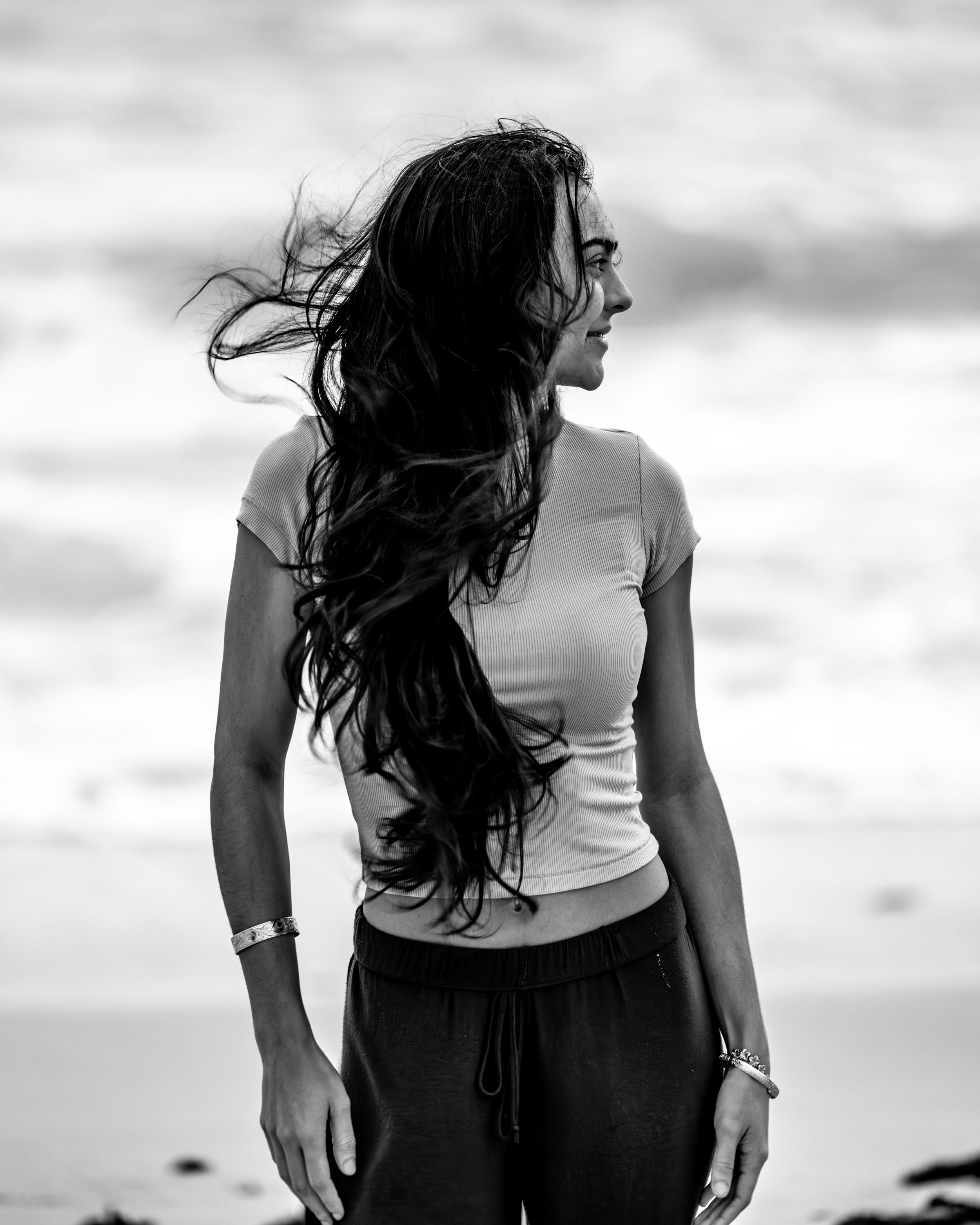 A black and white photo of a woman with long windswept hair standing by the ocean.