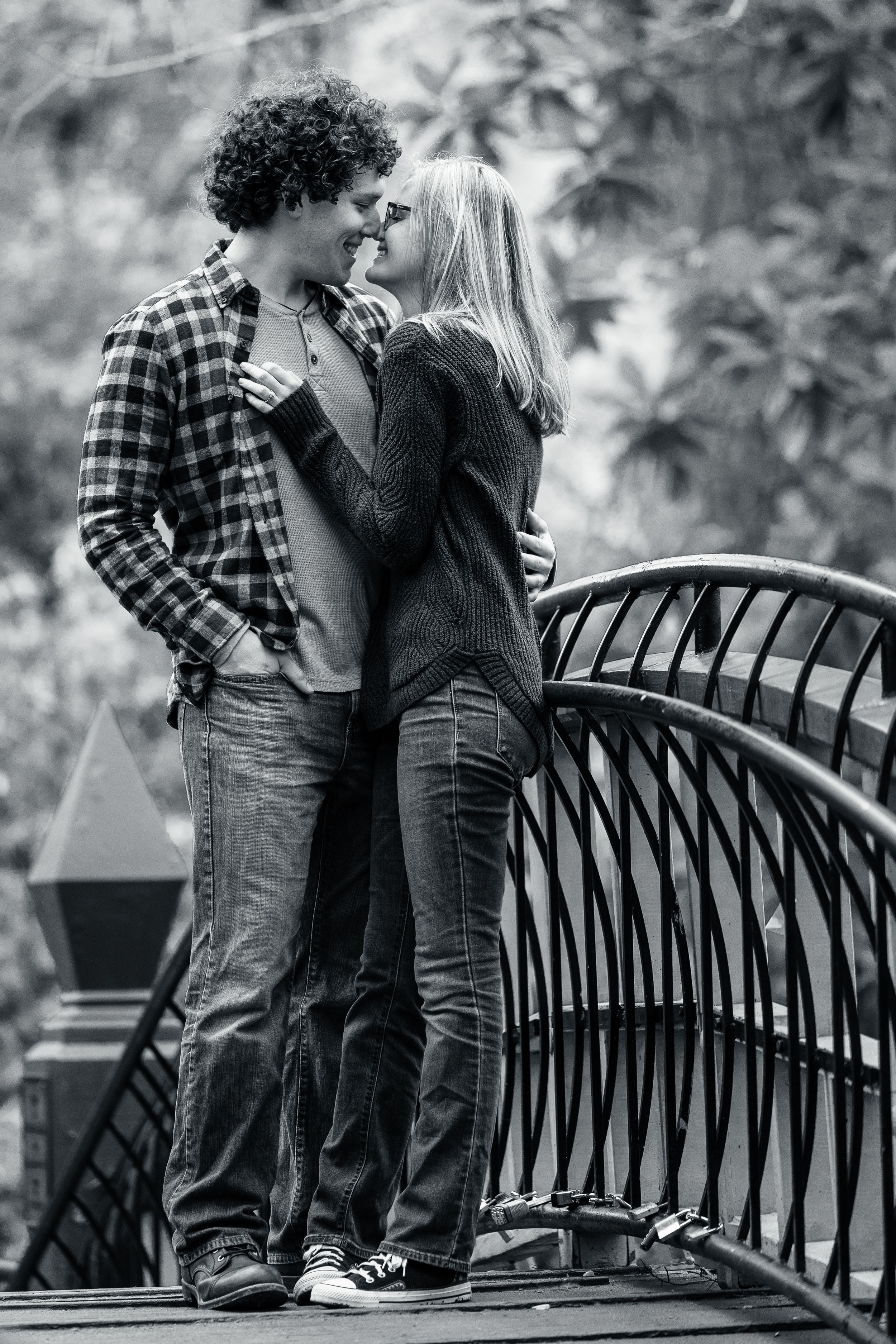 A black and white photo of a romantic couple almost kissing on a decorative bridge outdoors.