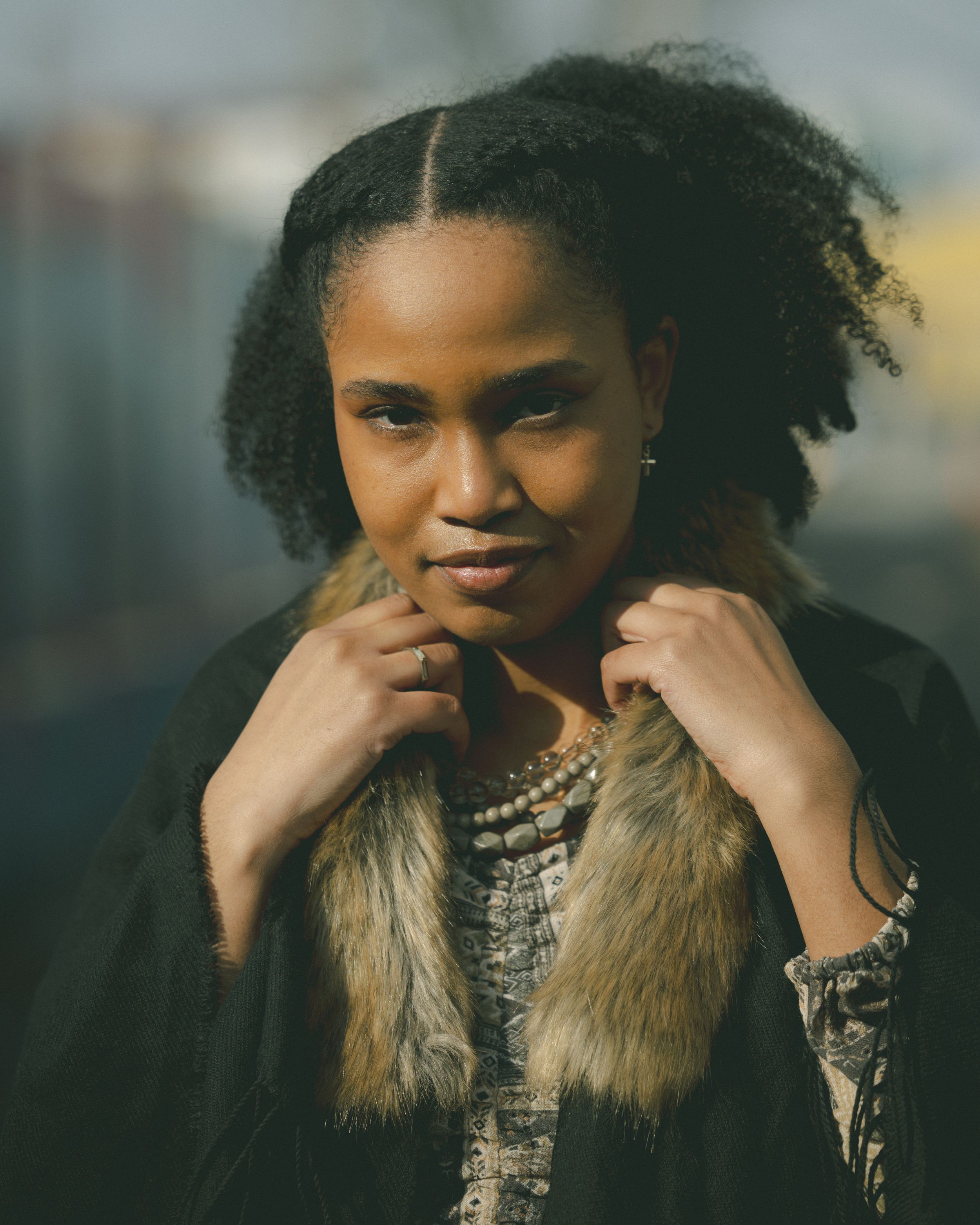 A young Black woman with natural hair holds her fur-trimmed coat collar in soft outdoor light.