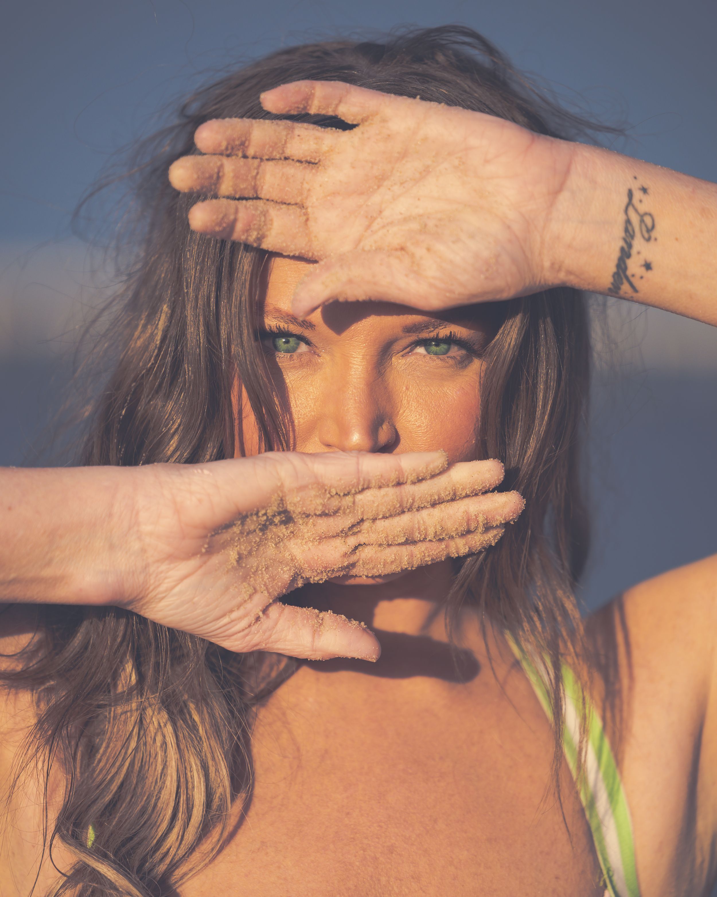 Woman with sandy hands covering face at beach in warm sunlight.