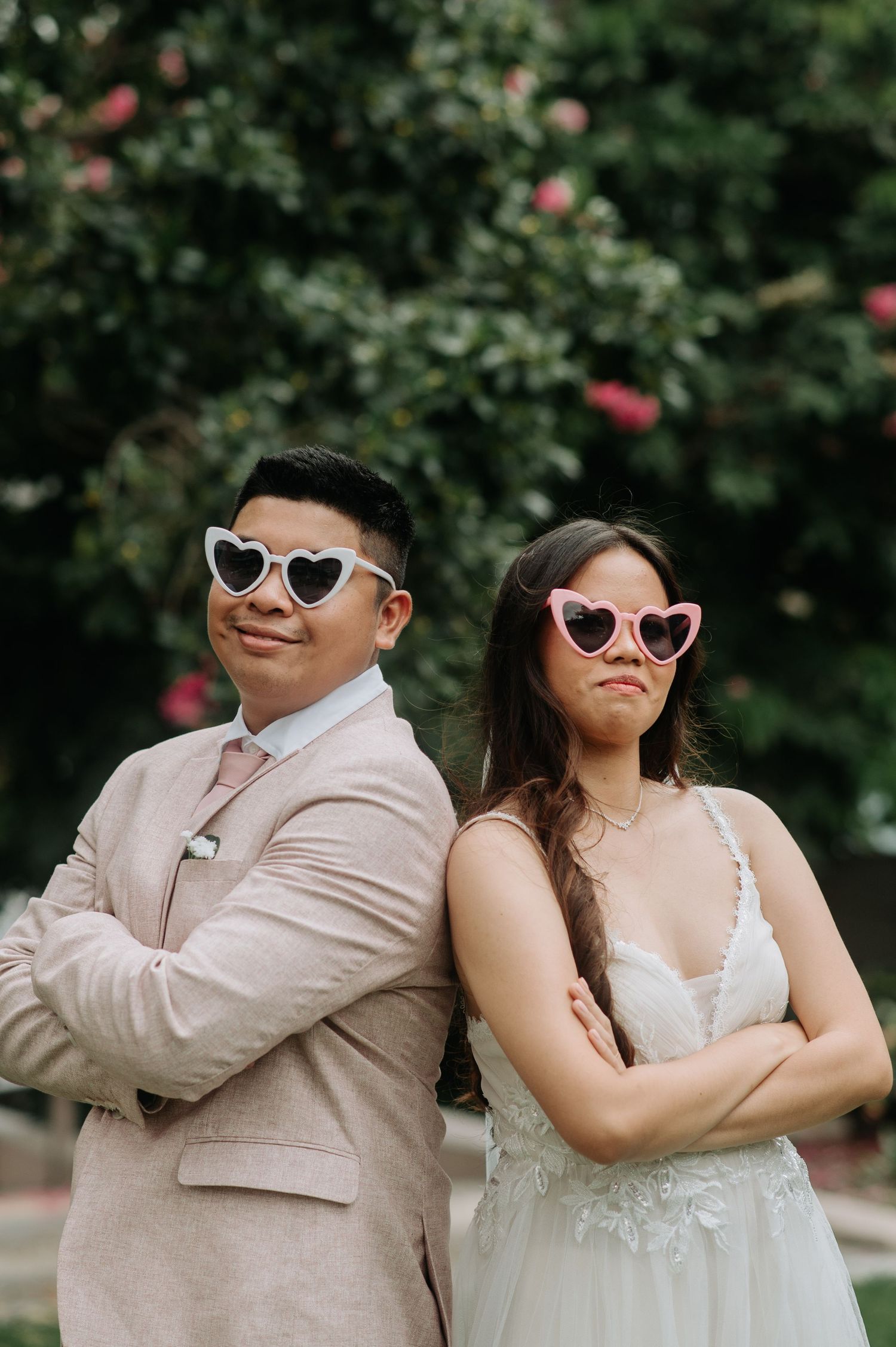 Playful wedding couple wearing heart-shaped sunglasses posing back-to-back in a garden setting.