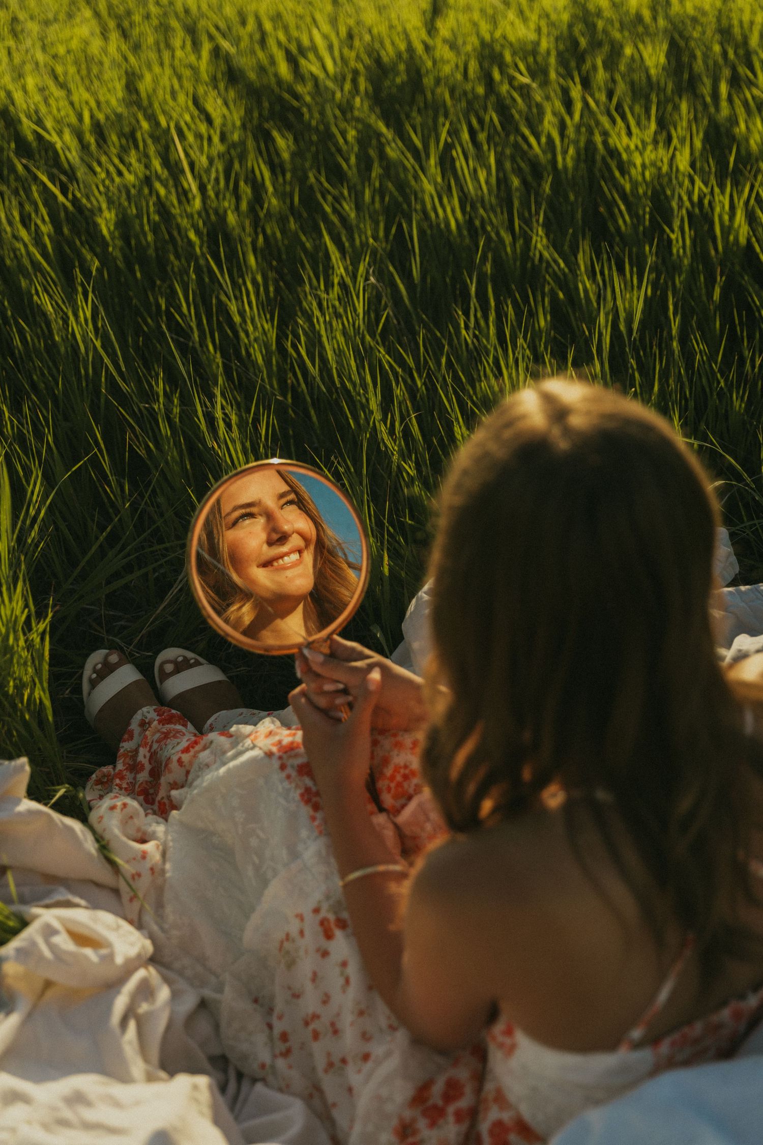Woman in white floral dress holding small round mirror while sitting in tall grass at sunset.
