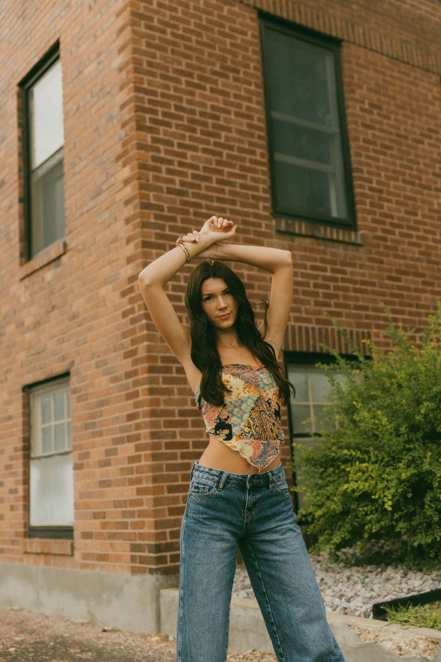 Woman in crop top and jeans posing against brick building exterior.