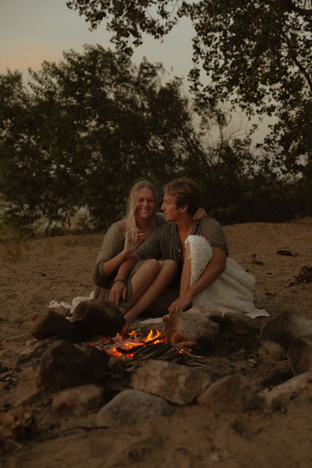 Two people relax by a cozy campfire on a rocky beach at dusk.