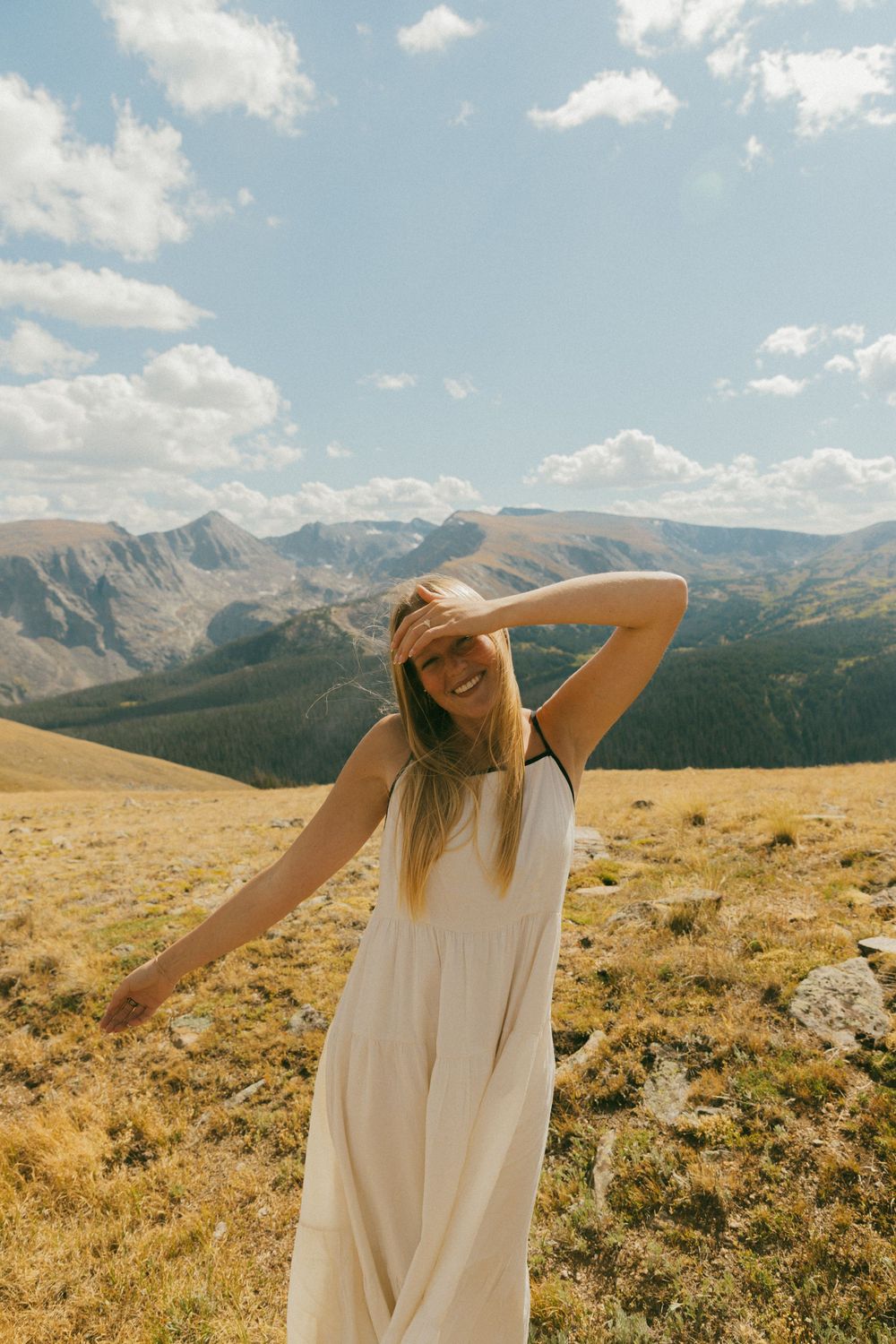Woman in white dress stretches her arms against a backdrop of mountain vistas.