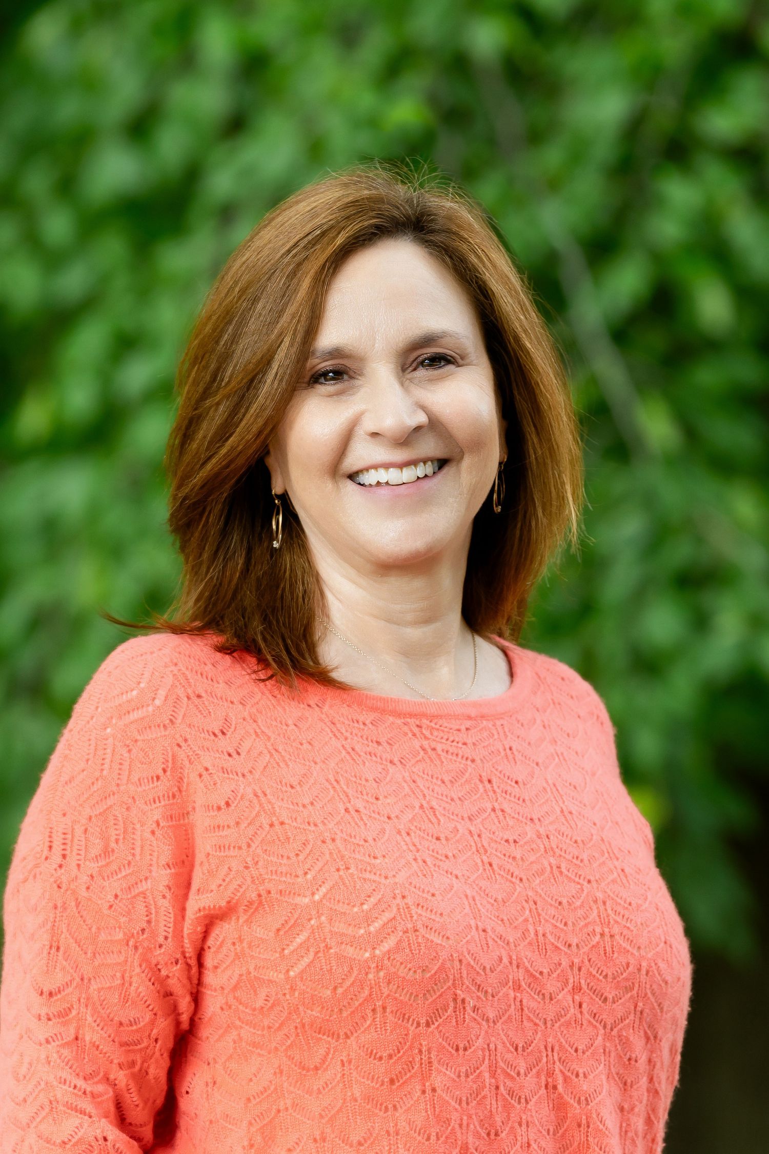 Portrait series featuring warm smile and coral textured sweater against natural green background.