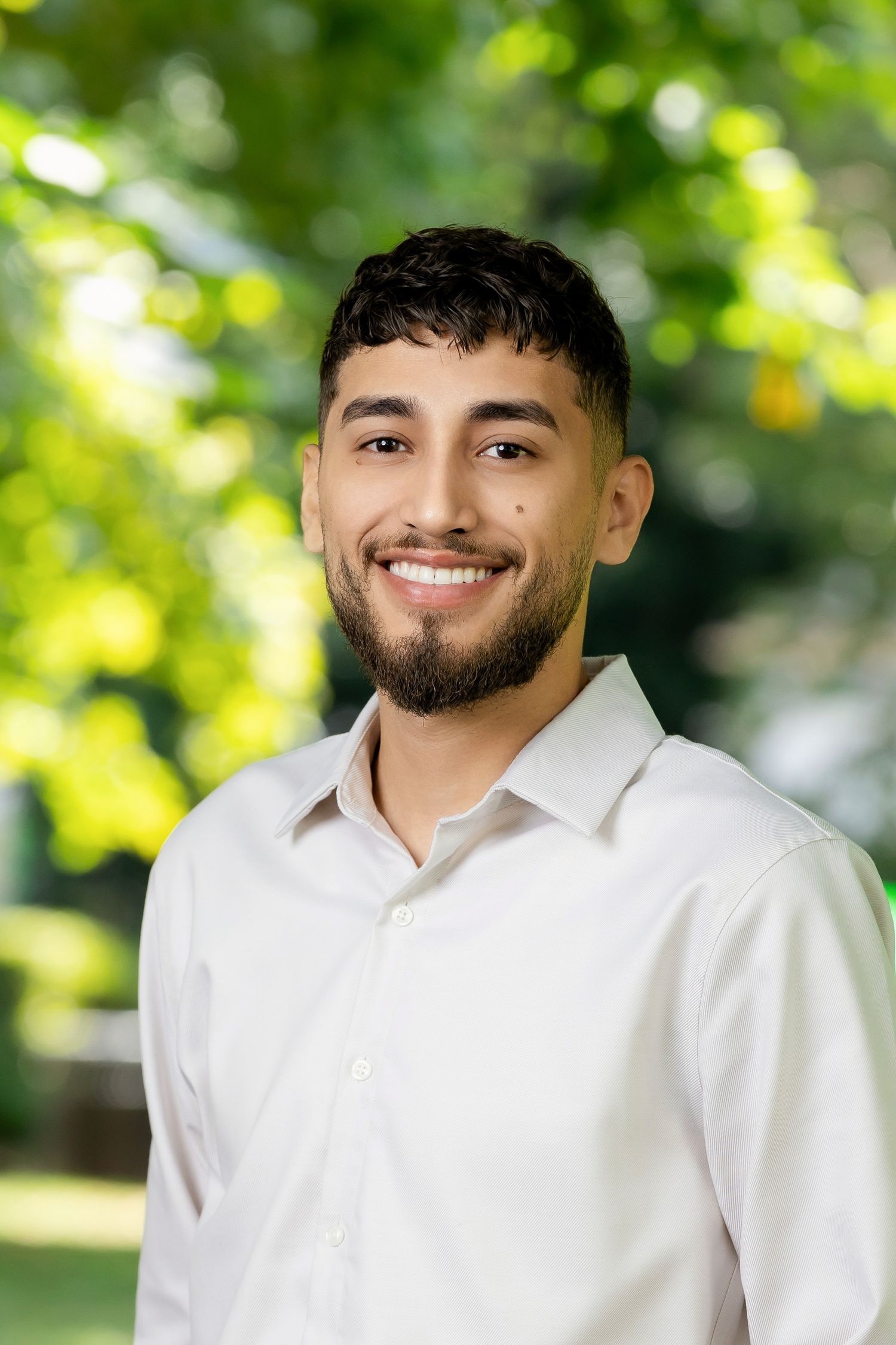 Portrait series showing white collared shirt and neat beard against outdoor greenery.