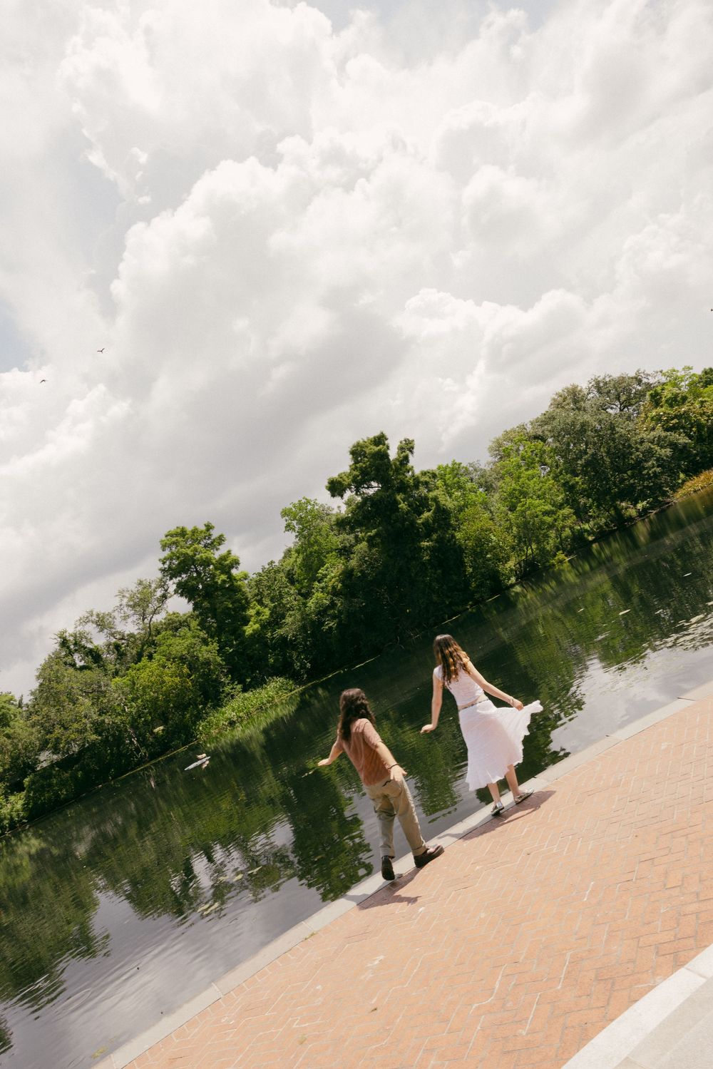 Two people in white dresses walk along a tranquil riverbank with trees and clouds reflected in the water.