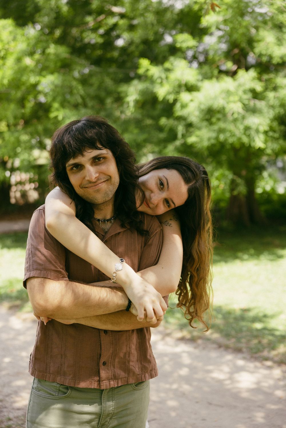 Two people share a warm embrace outdoors on a sunny day with lush green foliage in the background.