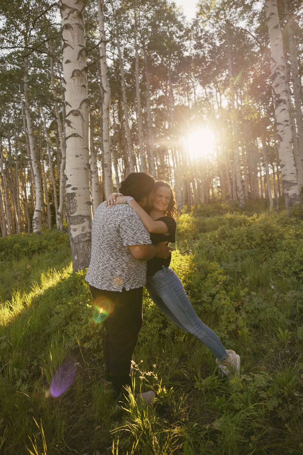 A couple embraces in a sun-dappled birch forest during sunset.