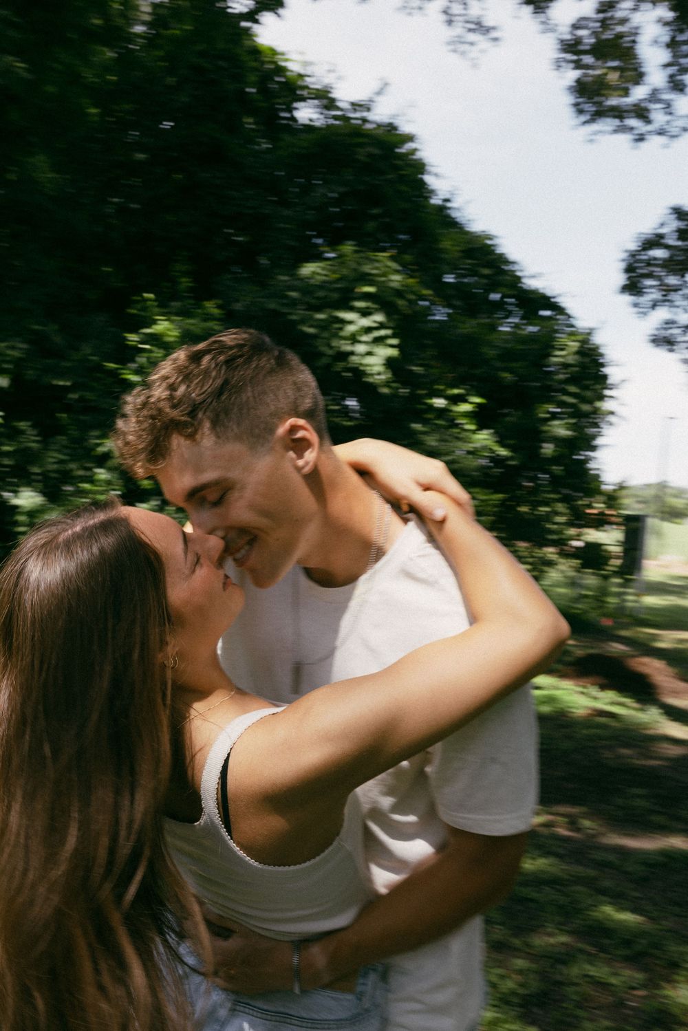 A romantic embrace captured outdoors with trees and natural lighting in the background.