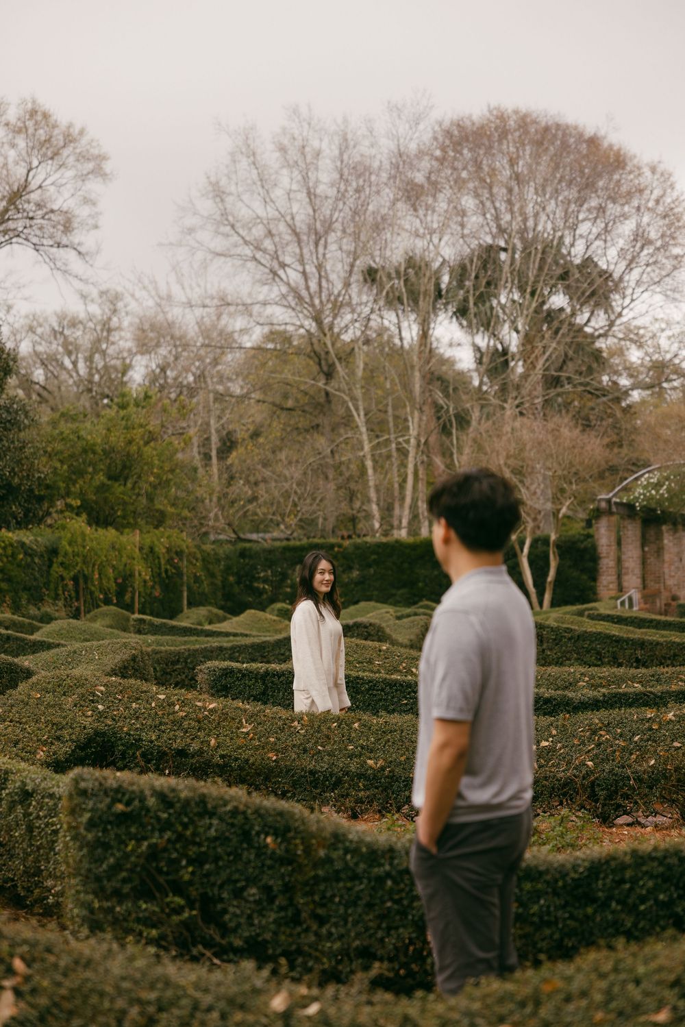 Two people share a moment while standing between rows of manicured hedges in a formal garden on an overcast day.