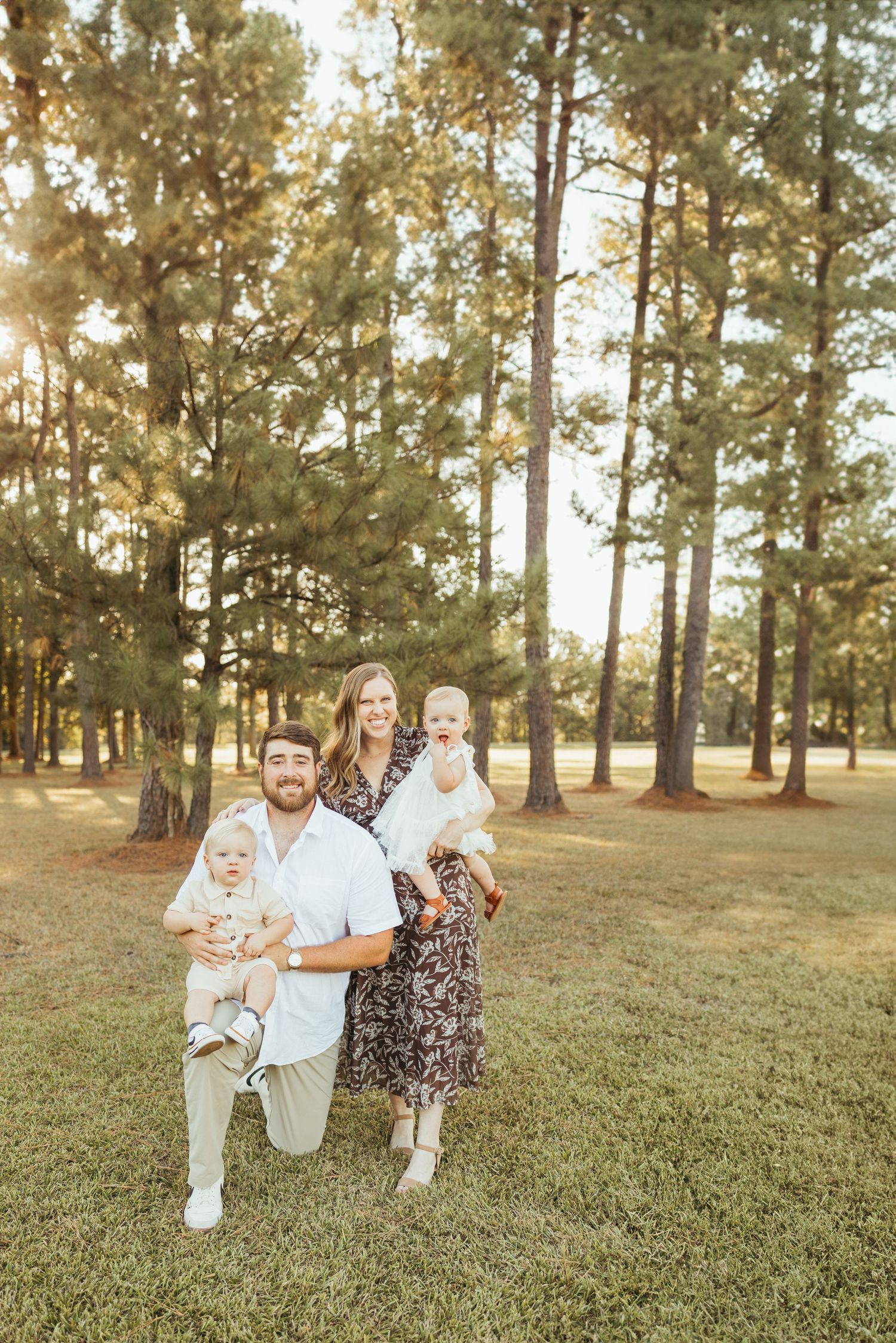 Family of four posing outdoors in pine forest, parents holding two babies in golden sunlight.