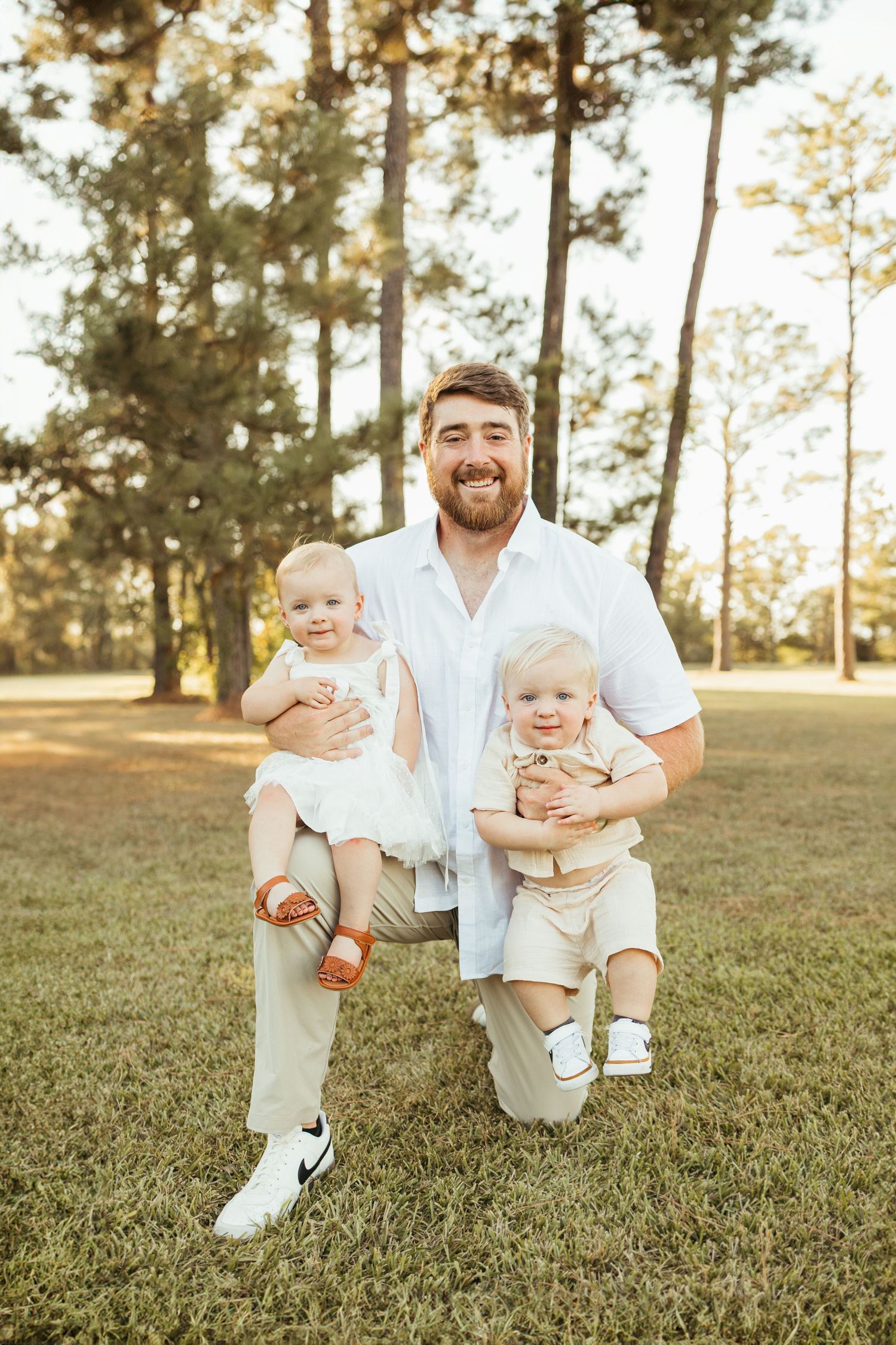 Family of four posing outdoors in pine forest, parents holding two babies in golden sunlight in Friendswood TX.