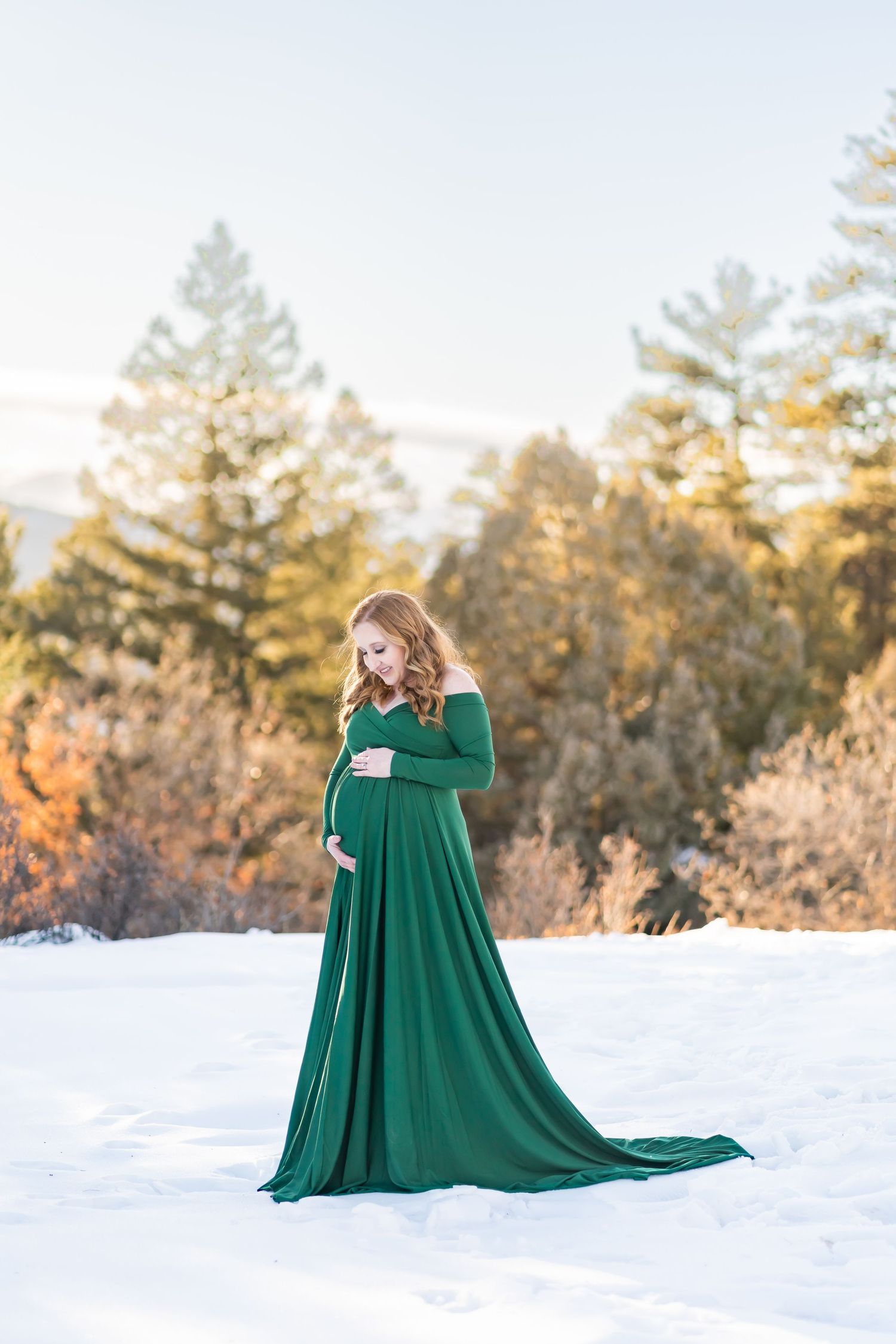 Winter maternity portrait in flowing emerald dress against snowy landscape with golden sunlight.