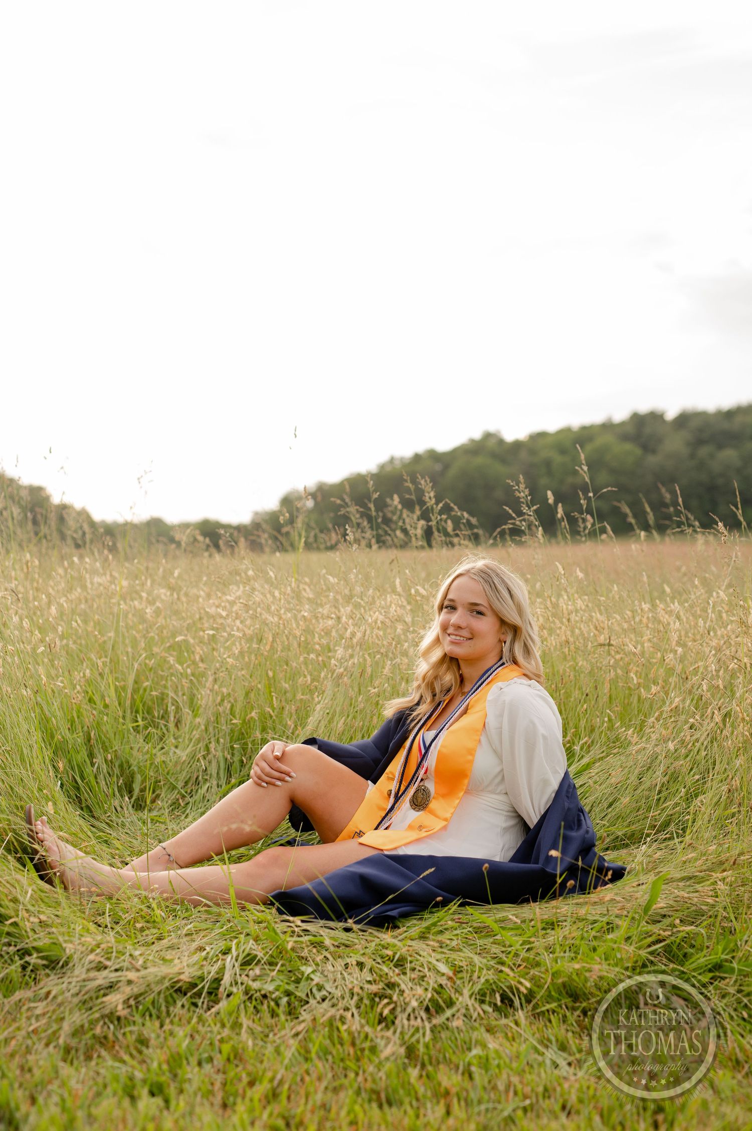 Graduate in casual attire sits among tall grass in a scenic meadow during photo session.