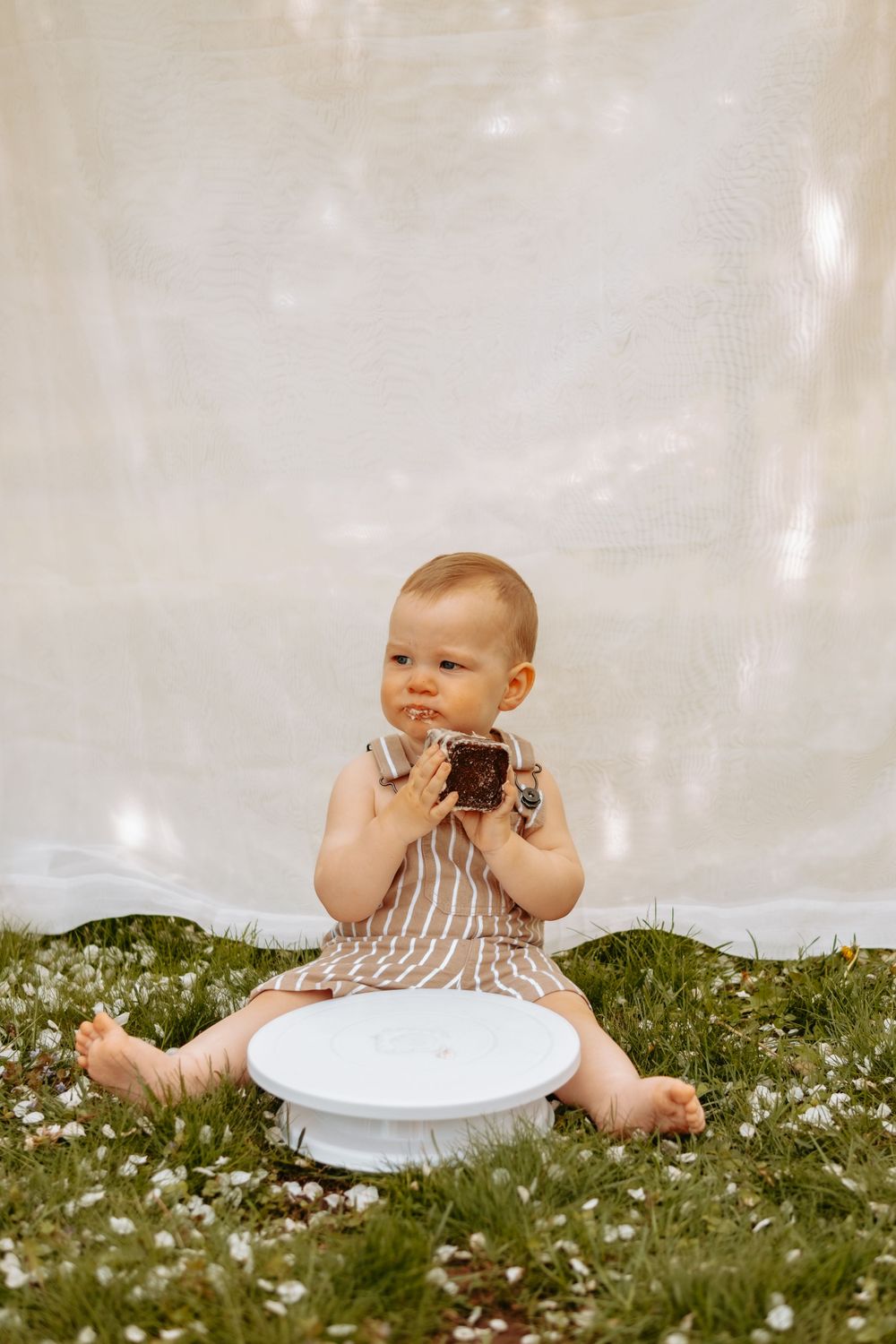 Small child in striped outfit eating chocolate cake outdoors against white backdrop.