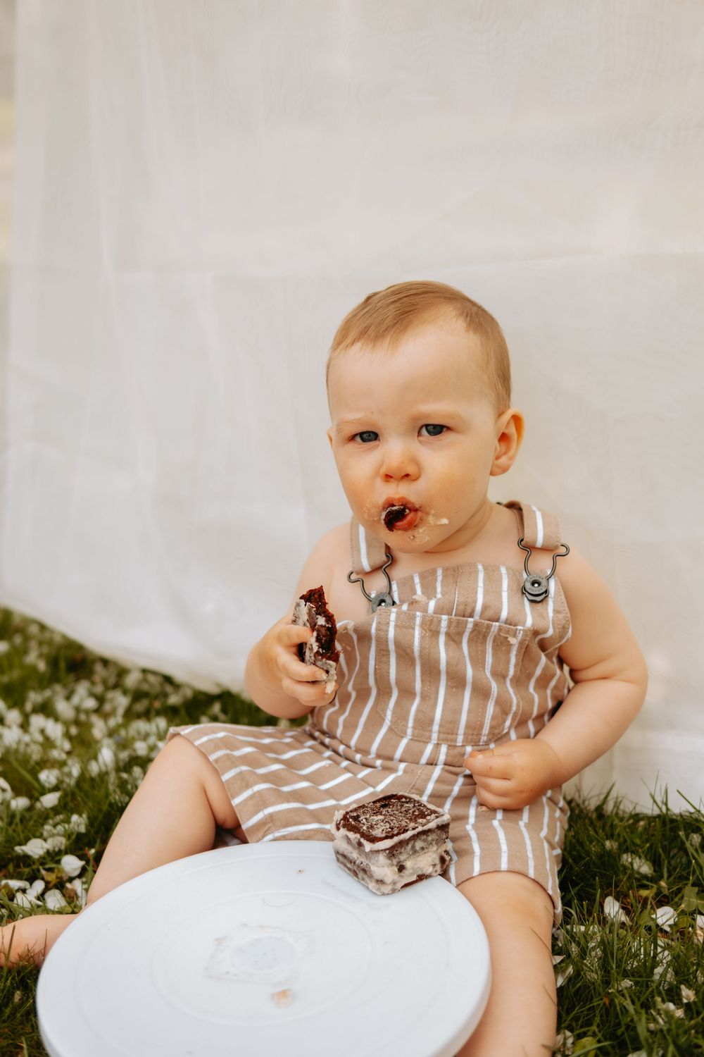 Baby wearing striped overalls enjoying messy cake smash on white plate outdoors.