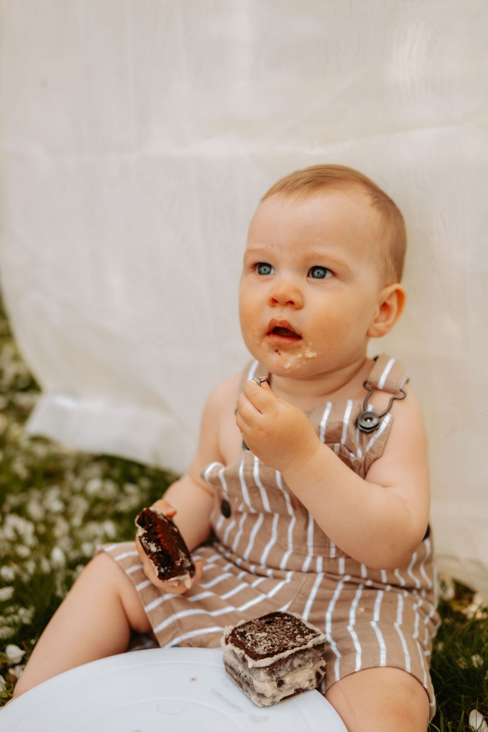 Baby in striped romper enjoying ice cream sandwiches while sitting on a white surface outdoors.