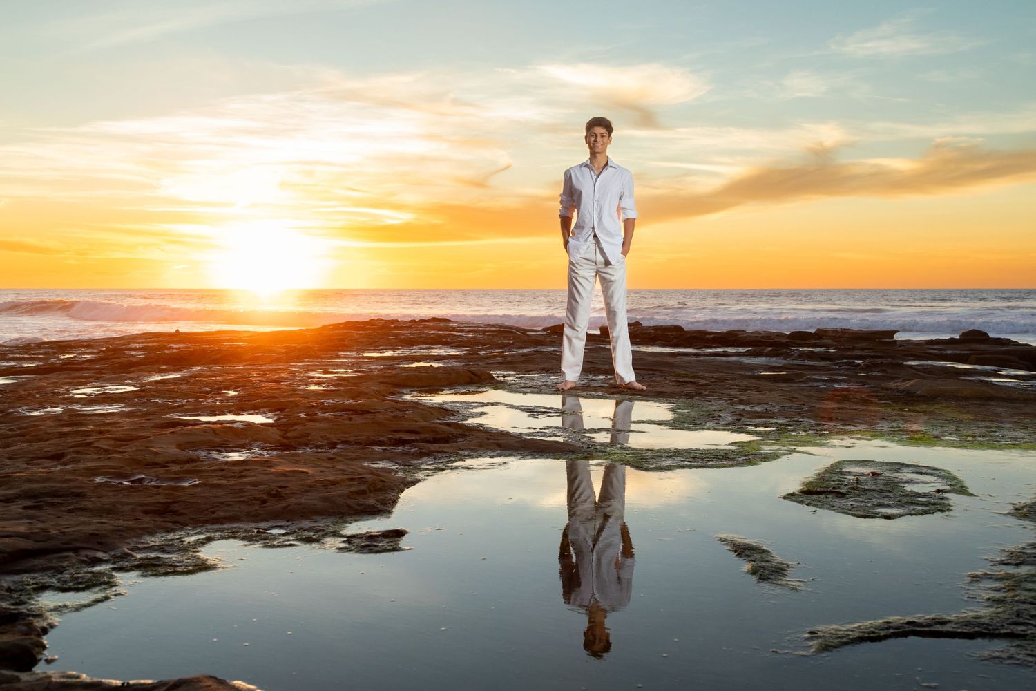 A silhouetted high school senior in white clothing stands on a reflective tide pool by the ocean during a golden sunset.