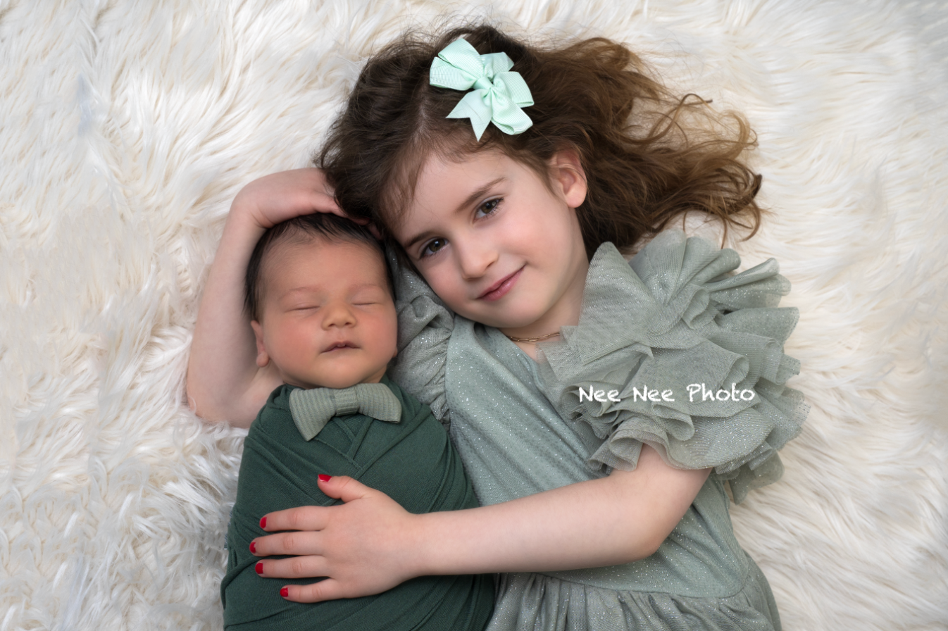 Older sister in sage green dress gently holds sleeping newborn wrapped in matching green swaddle on white fur.