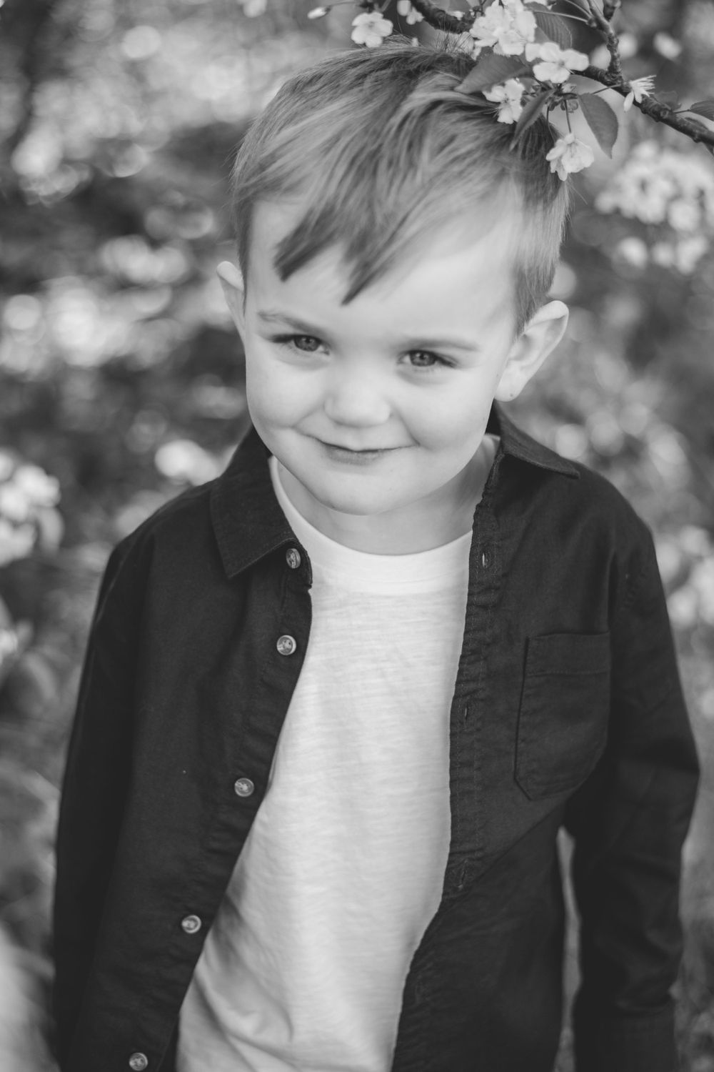 Black and white portrait of young child in button-up shirt standing by spring blossoms outdoors.