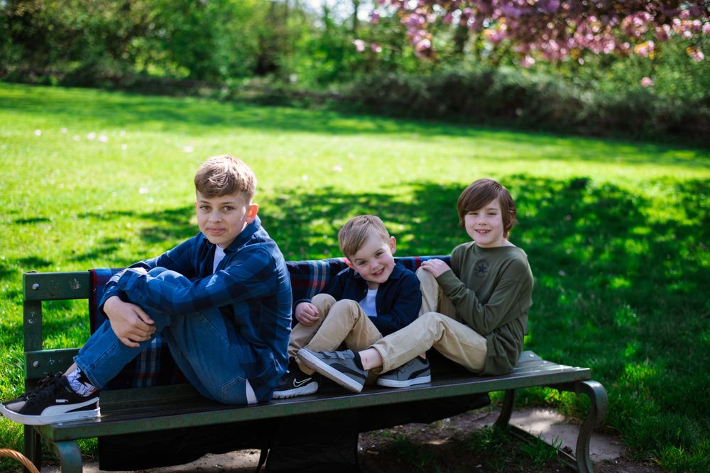Three young people sitting casually on a wooden bench in a sunny garden.