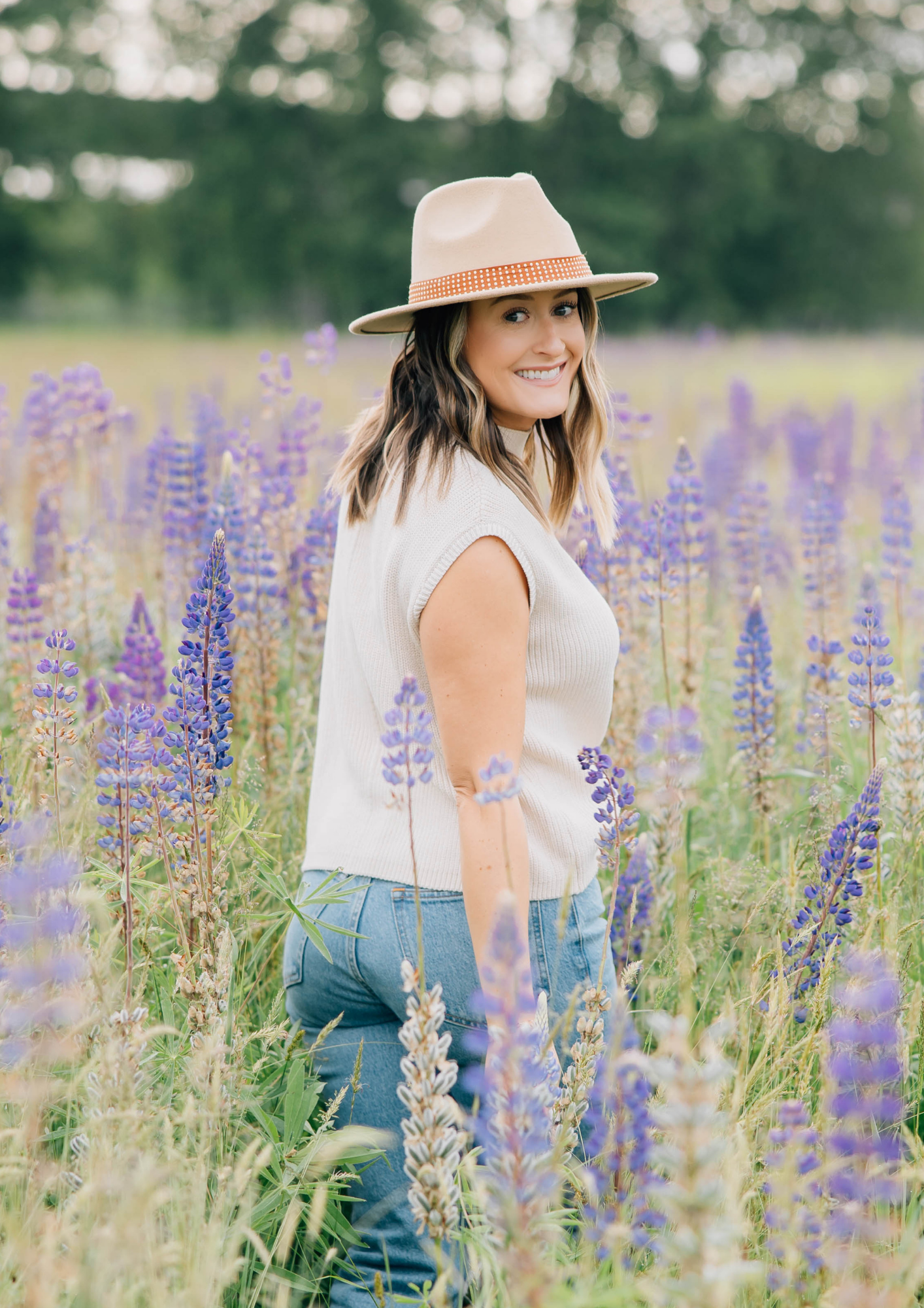 Woman in beige hat and white top smiling in purple lupine flower field.