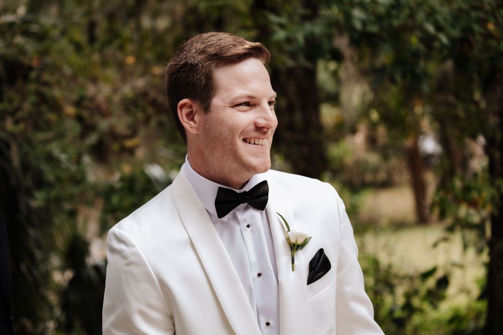Groom in classic white tuxedo and black bow tie smiling outdoors. wedding at la venture grove in green cove springs Florida