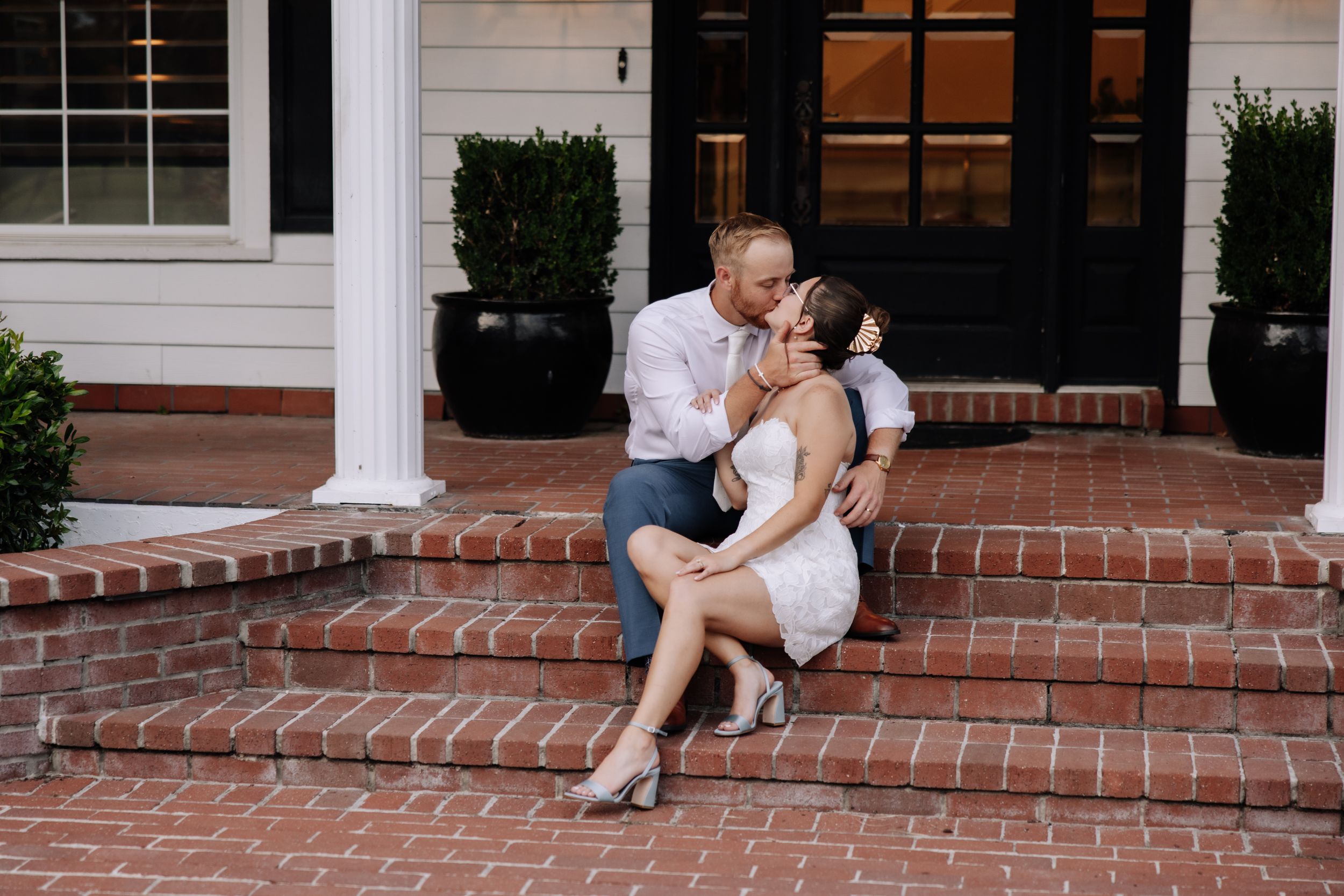 Couple sharing a romantic kiss on brick steps outside a white colonial home during an engagement photo session.