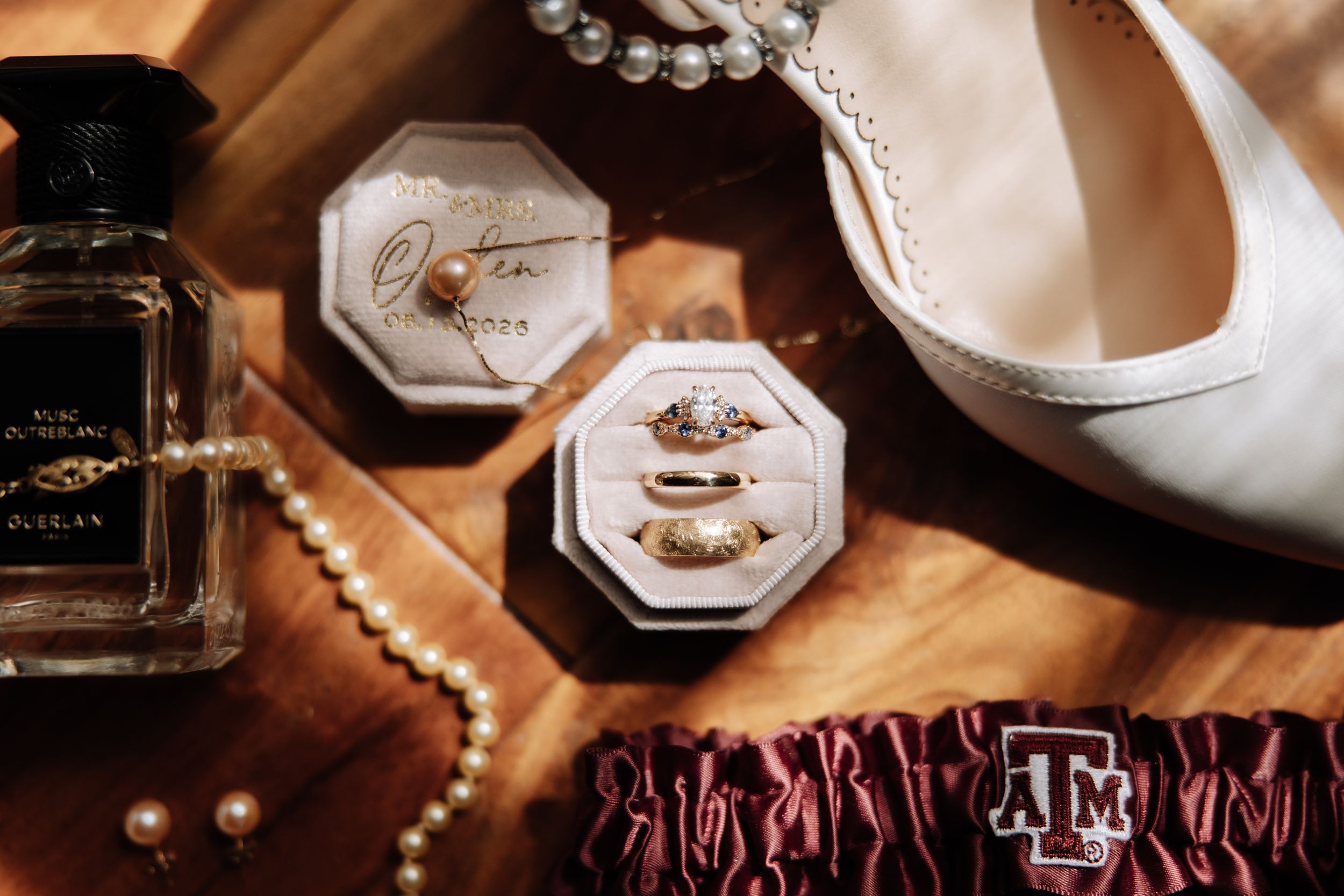 Wedding flat lay with diamond rings in velvet box, pearl necklace, perfume bottle, and white bridal heels on wood surface.
