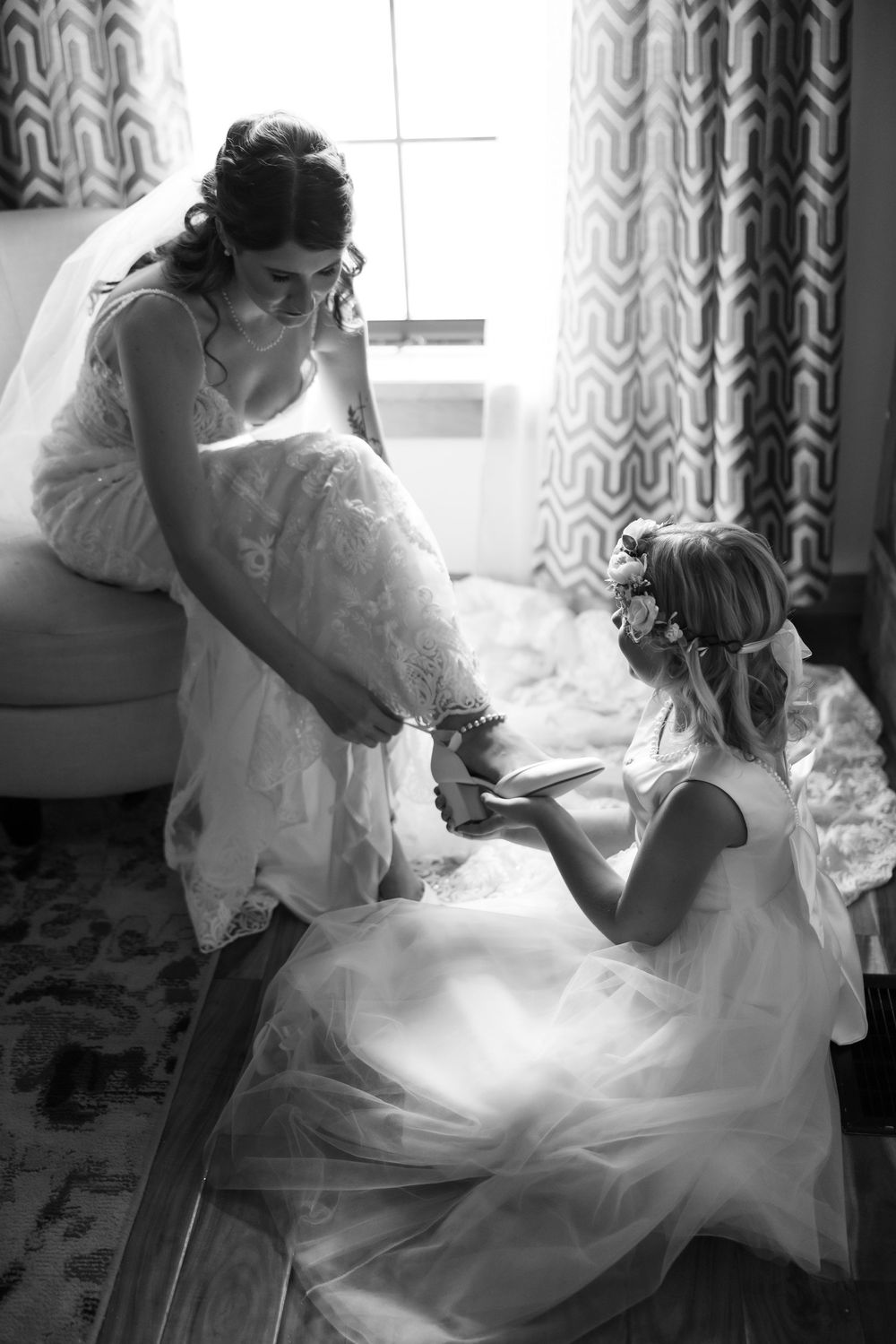 Black and white photo of a bride having her shoes put on by a flower girl in a floral crown before the wedding ceremony.