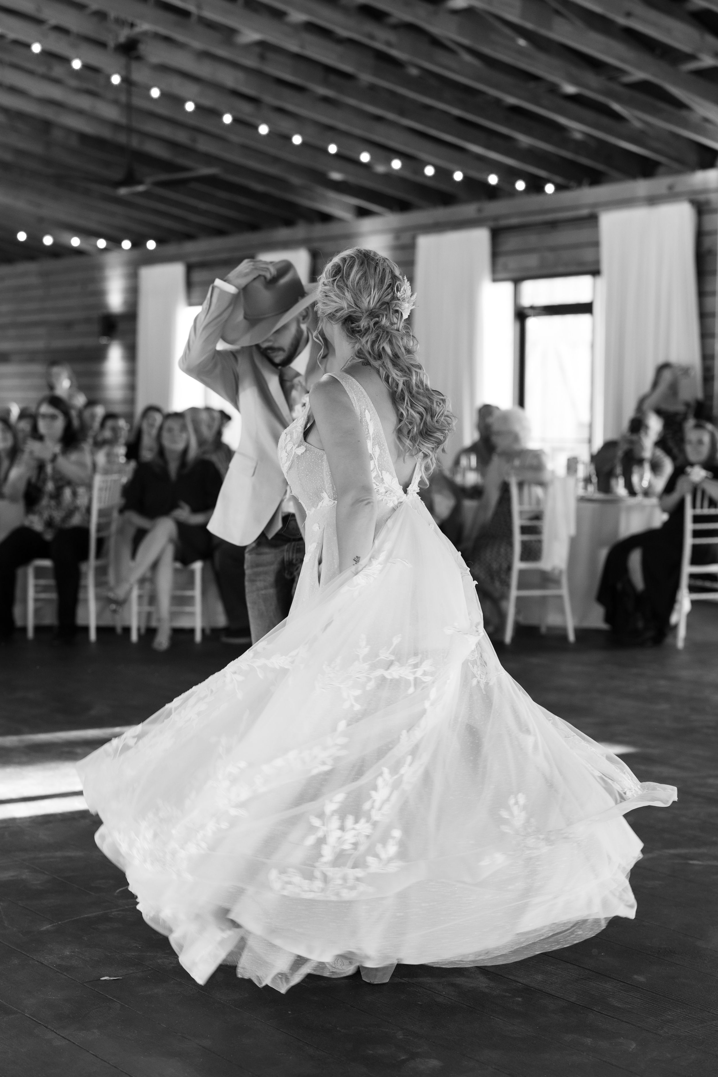 Black and white photo of bride and groom sharing first dance in elegant barn venue with string lights.