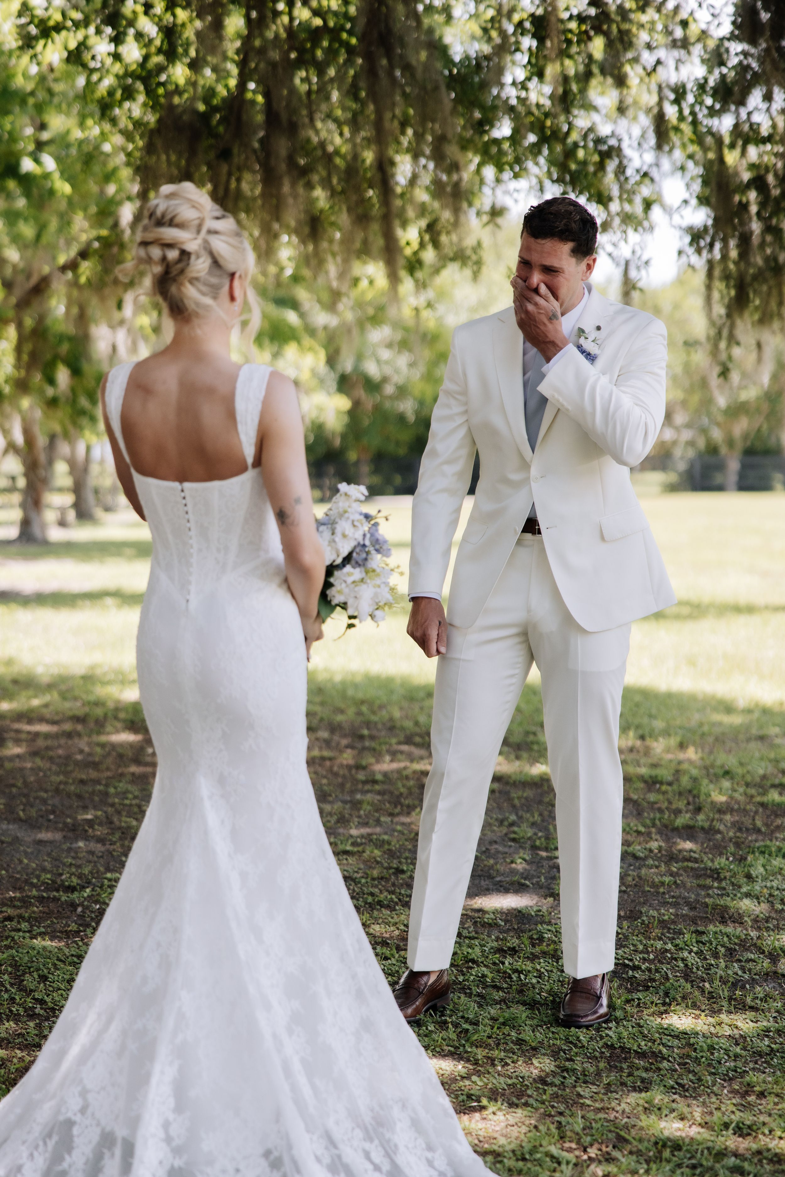 Emotional groom in white suit covering his mouth as he sees his bride in a lace wedding dress for the first time outdoors.