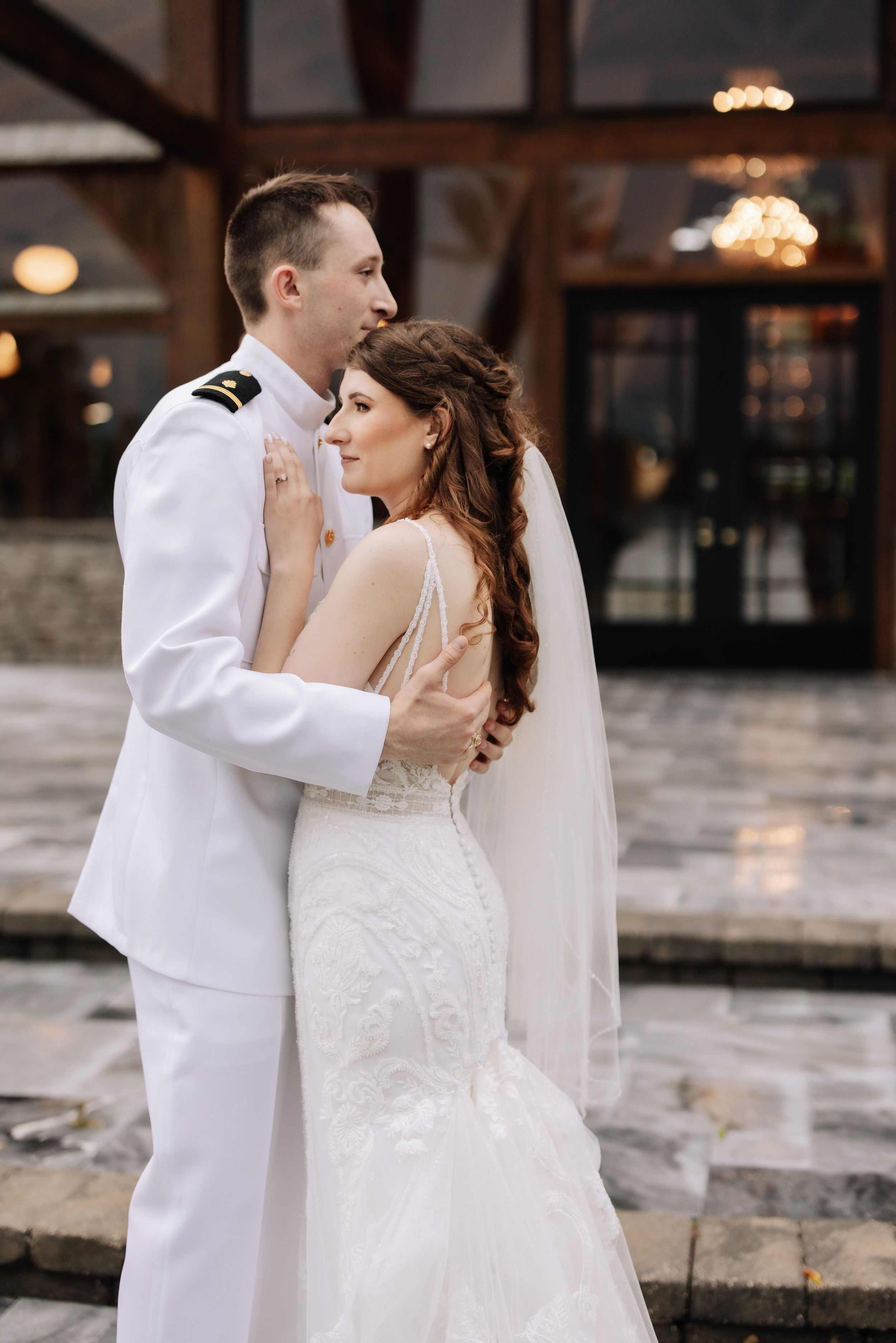 A bride in a lace gown and veil embraces a groom in a white naval uniform outdoors at their wedding.