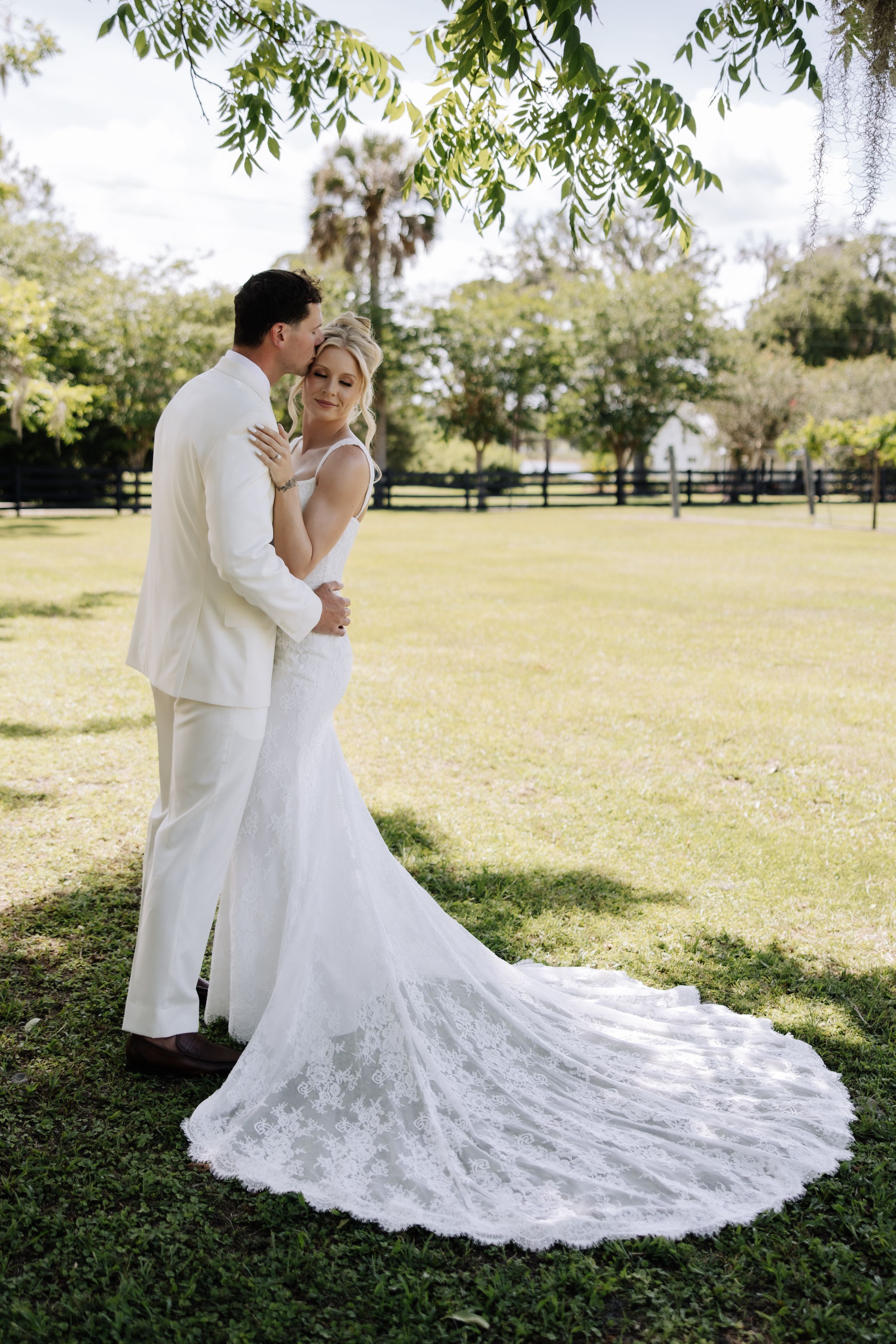 Bride and groom embrace on lush green lawn, bride wearing elegant lace wedding gown with long train.