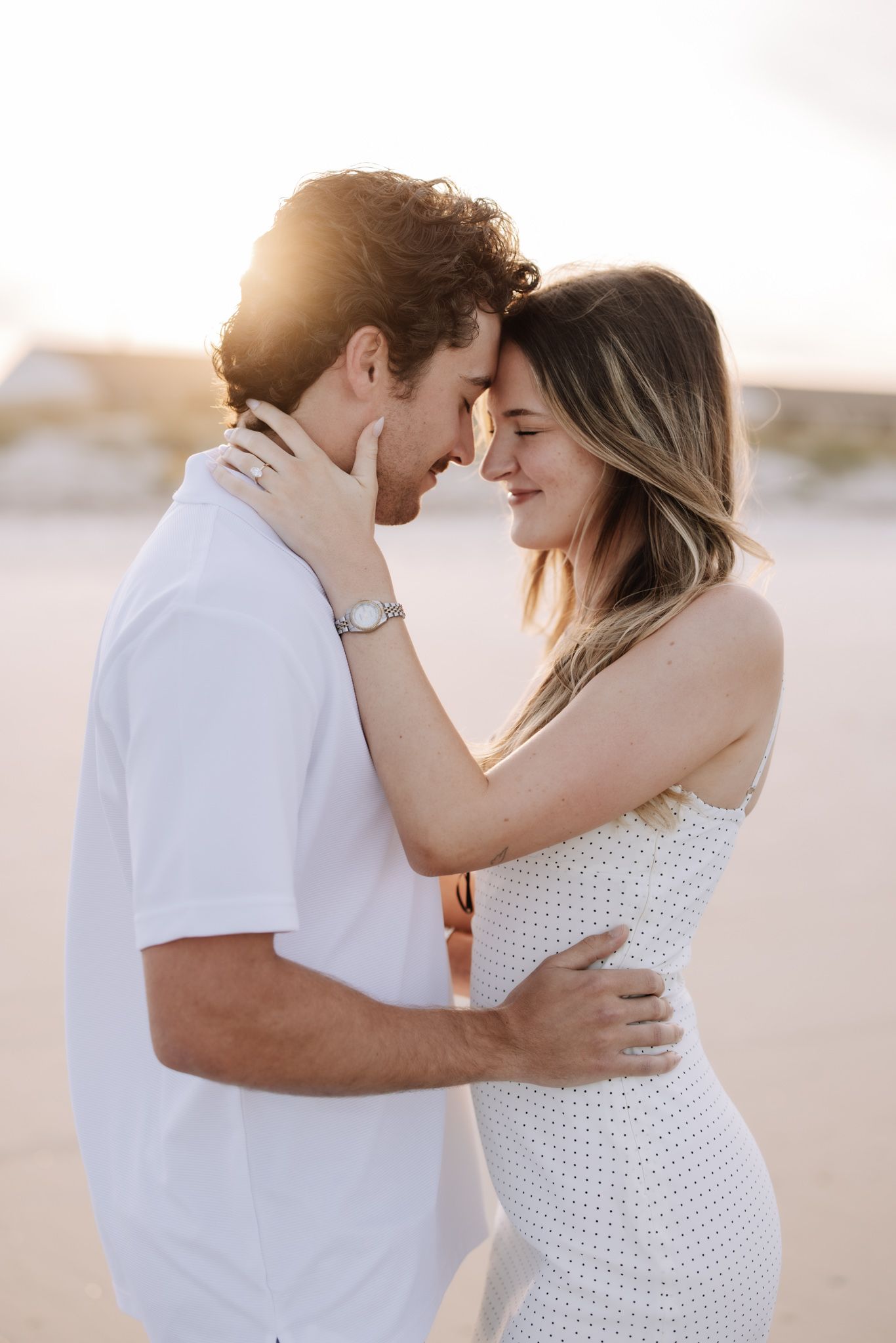 Couple sharing a romantic forehead touch at sunset on a beach, dressed in white outfits.