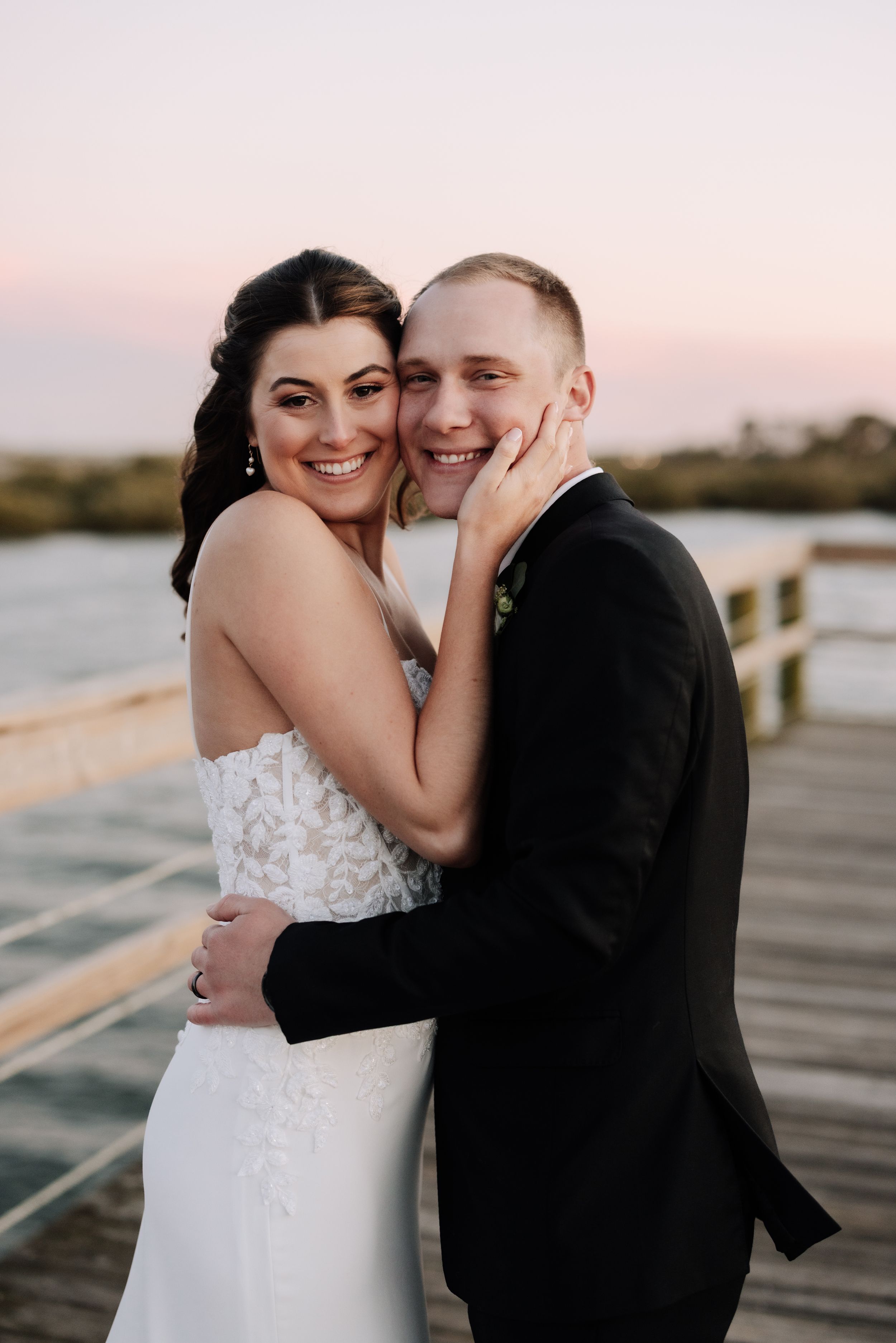 A bride and groom embrace on a waterfront pier at sunset, smiling joyfully in wedding attire.