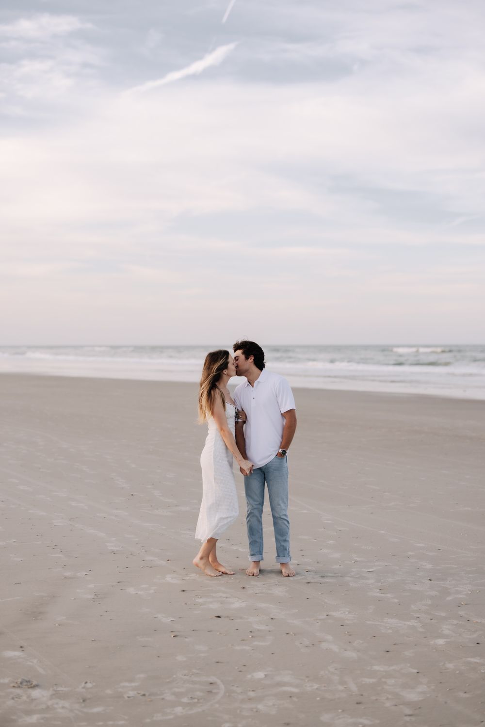 Couple sharing a romantic kiss on a sandy beach, dressed in white outfits with ocean waves in the background.