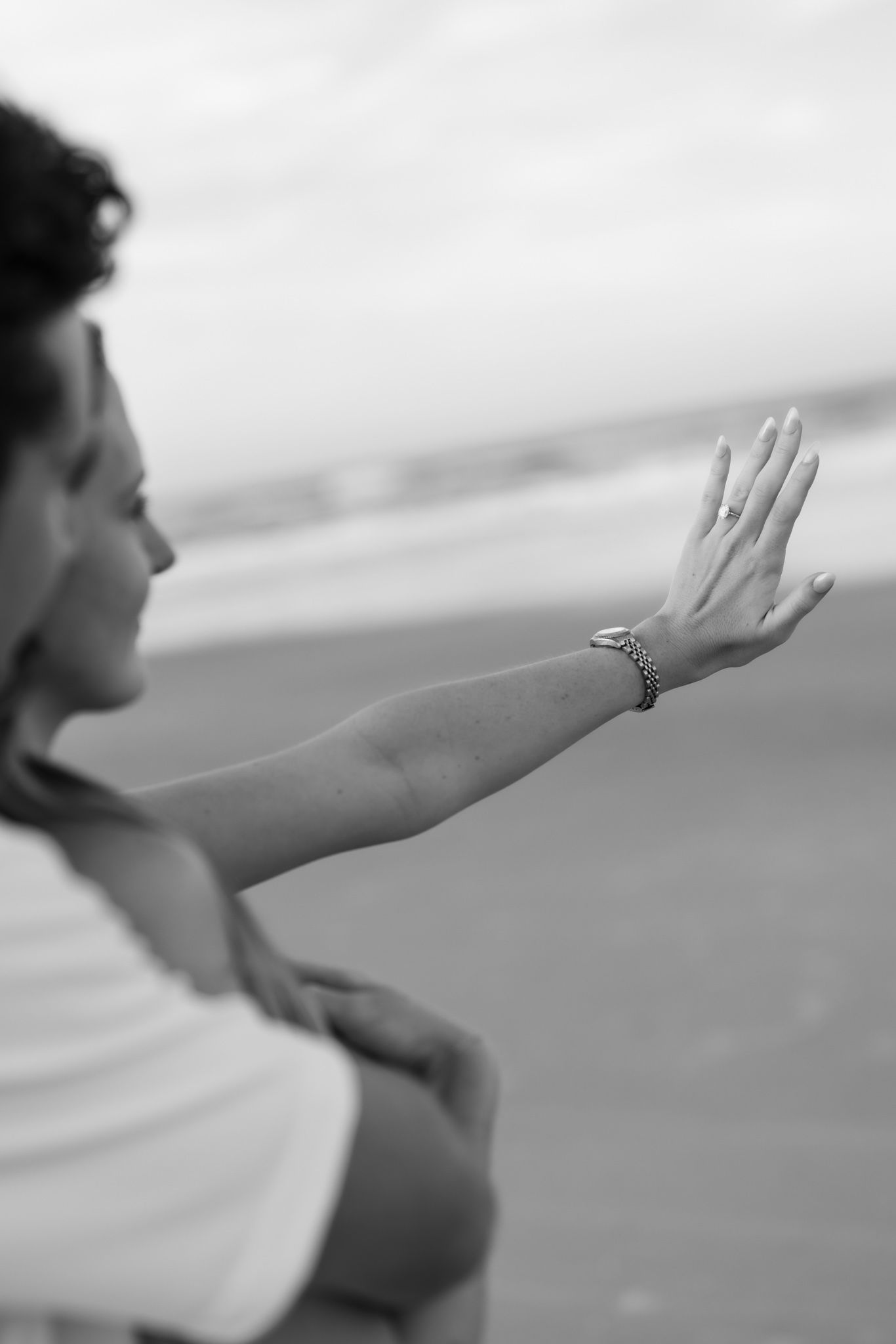 Black and white photo of a woman at the beach extending her hand showing a bracelet, with ocean waves in the background.