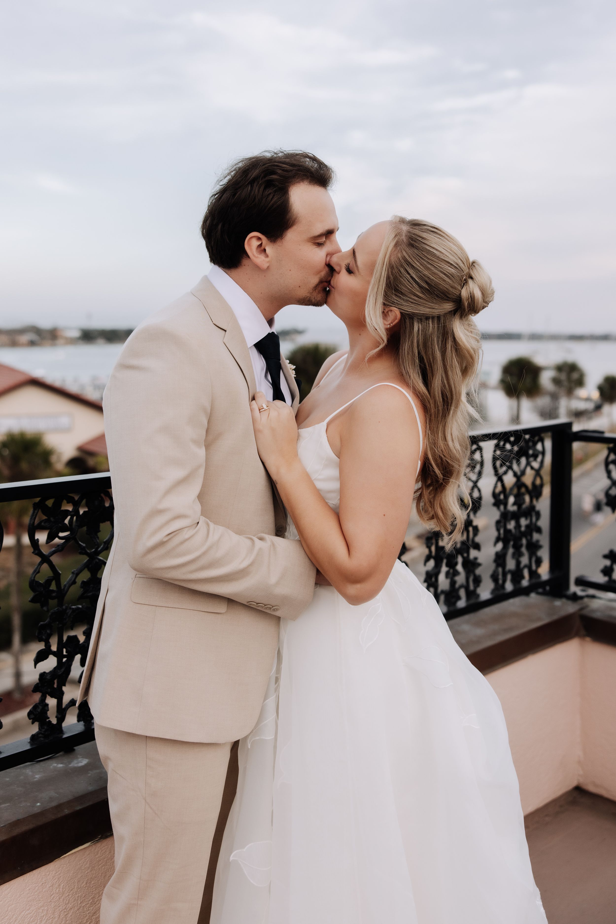 A bride and groom share a romantic kiss on a balcony overlooking the water, she in white gown, he in tan suit.